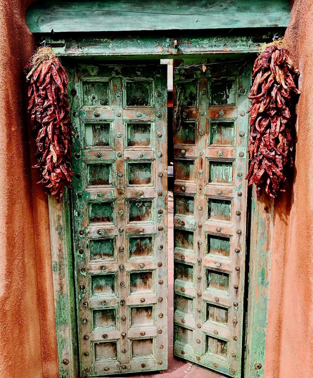 Weathered green wooden double doors with dried red chile ristras hanging on each side, adobe walls along Santa Fe, New Mexico streets