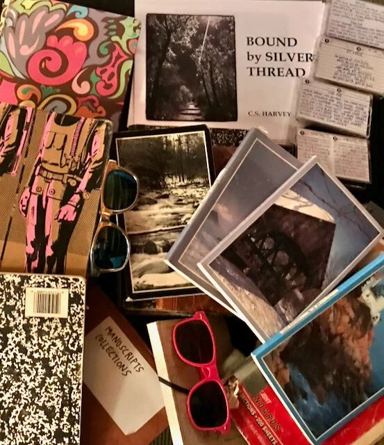 Photograph of a collection of notebooks, many featuring landscape photos on the cover, lying scattered over a folder labeled "Manuscripts Collections", a printed copy of Bound by Silver Thread, four mix cassette taps, and  two pairs of sunglasses.