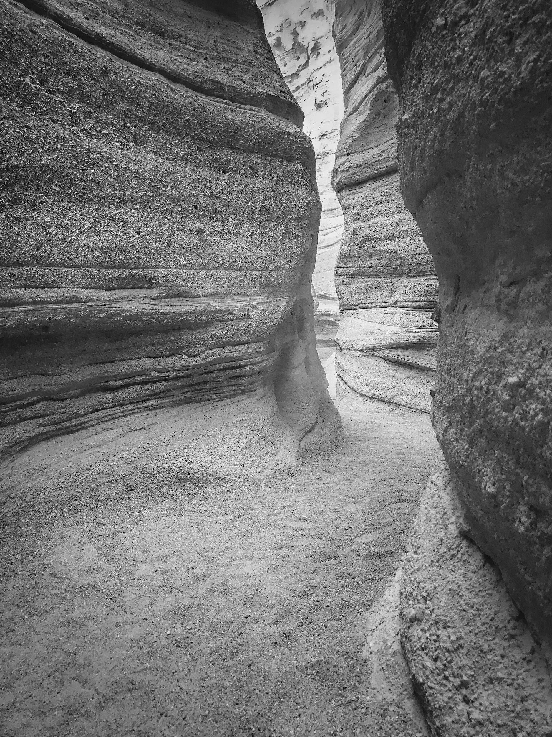 Black and white photo of narrow canyon walls with layered, textured rock formations, showing a winding trail through the slot canyon at Kasha-Kutuwe Tent Rocks in New Mexico.