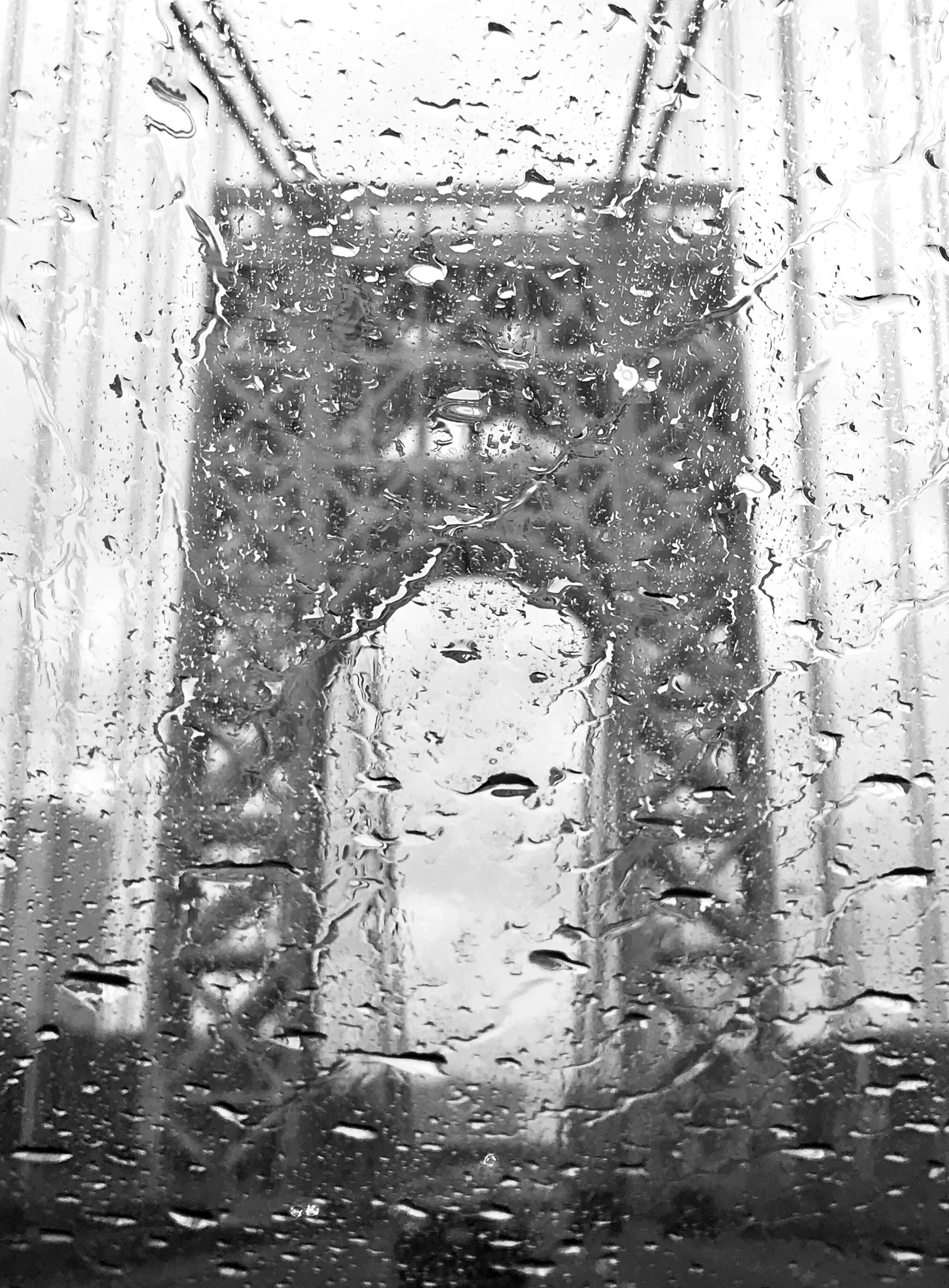 A black and white photograph taken through a rain-soaked windshield showing water lines and raindrops while in the background the outline of the George Washington Bridge rises up heading out of New York City
