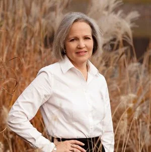 Portrait of a middle-aged woman with gray hair, wearing a white blouse, standing outdoors in a field of tall grasses.