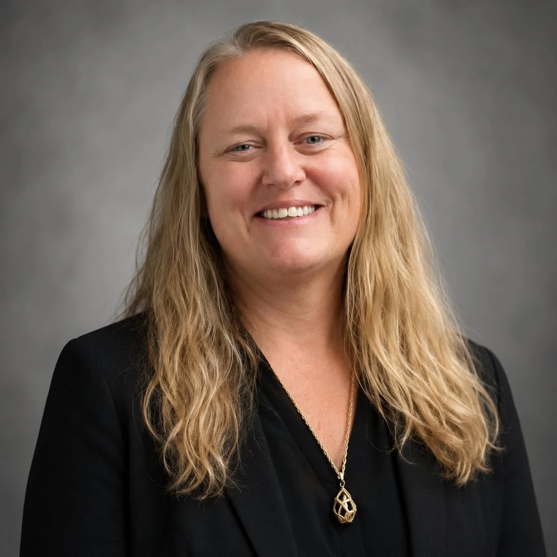 Professional portrait of a woman with long blonde hair, wearing a black blazer and gold necklace, smiling against a grey background.