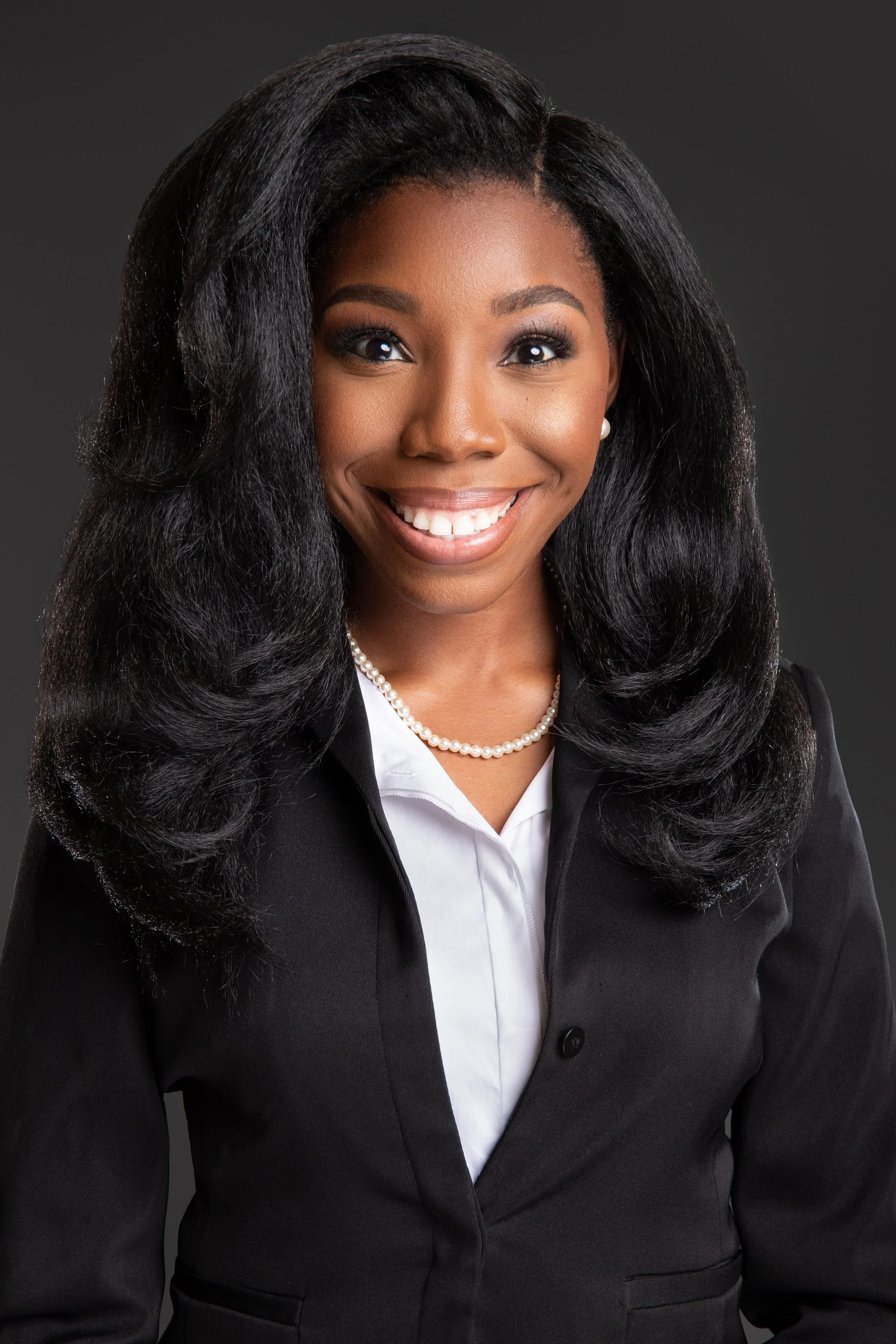 Professional portrait of an African American woman smiling, wearing a black blazer, white shirt, pearl necklace, and pearl earrings, against a dark gray background. professional black woman