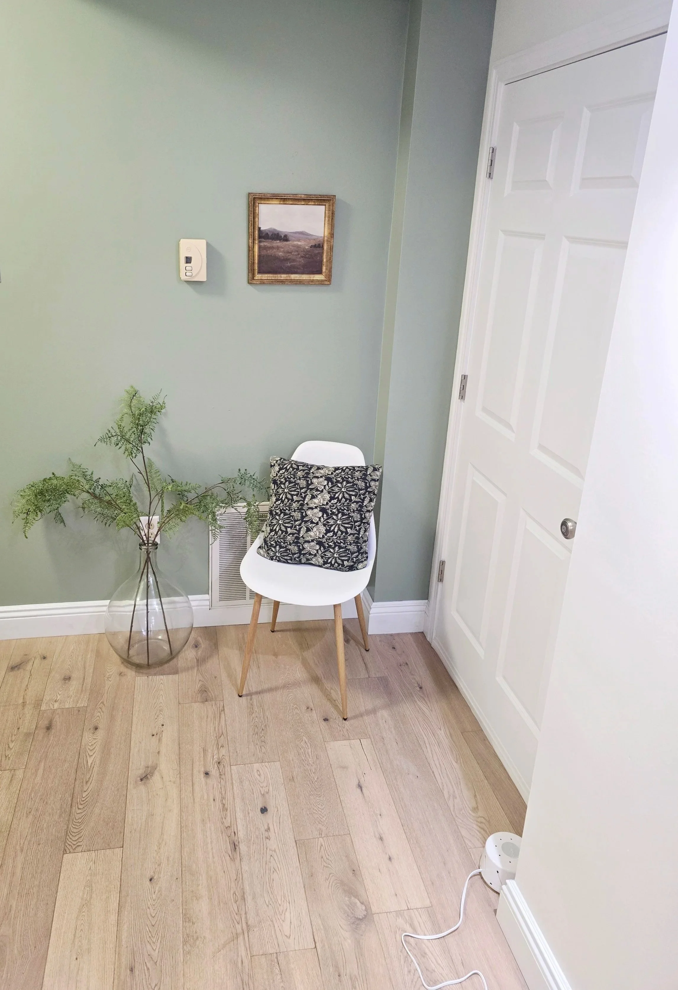 A corner of a room with light green walls, a white door, a white chair with a black and white patterned pillow, a glass vase with green fern leaves, a framed landscape painting, a wall-mounted thermostat, and a small white humidifier on the floor.