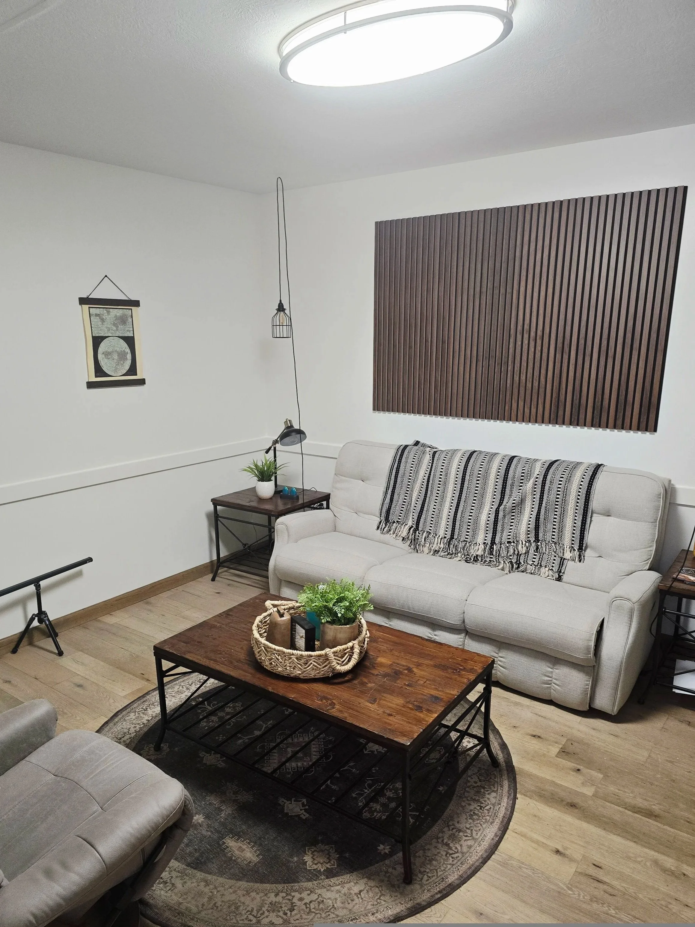 Living room with white sofa, wooden coffee table with a basket and plants, gray armchair, wall-mounted artwork, wooden wall decor, ceiling light, small table with lamp and plant, and wooden flooring.