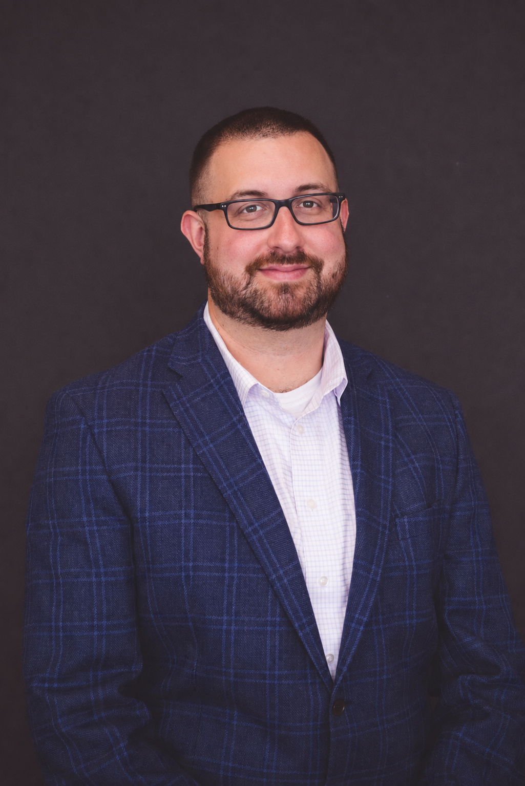 A man with glasses and a beard wearing a blue plaid blazer and a white shirt, posing against a dark background.