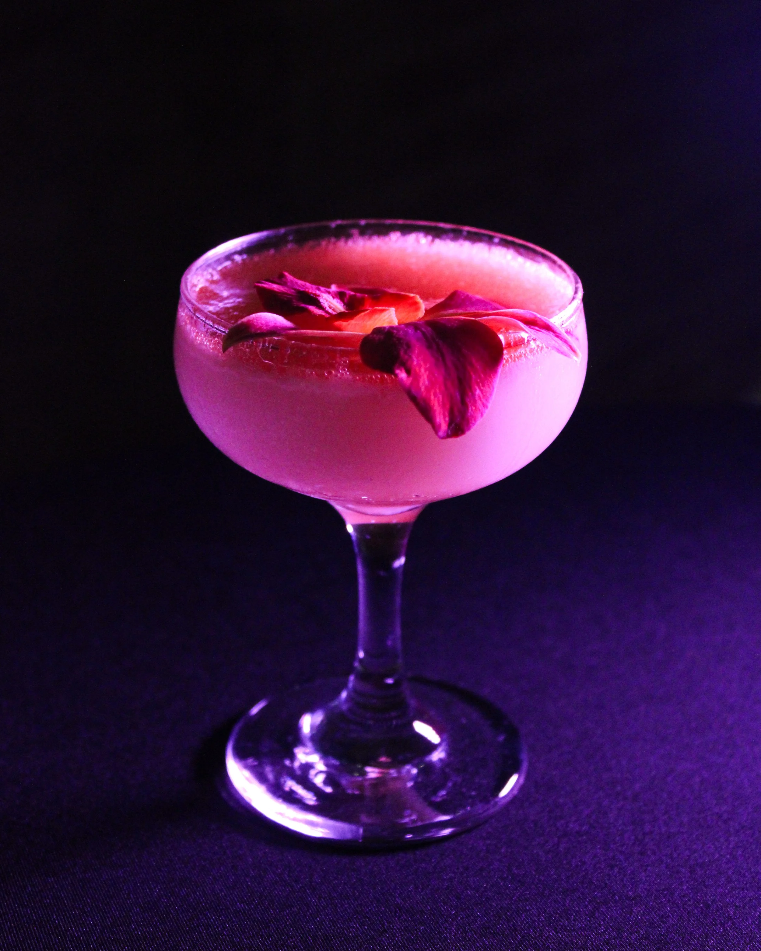 A pink cocktail in a coupe glass, garnished with flower petals, glowing under purple lighting against a black background.