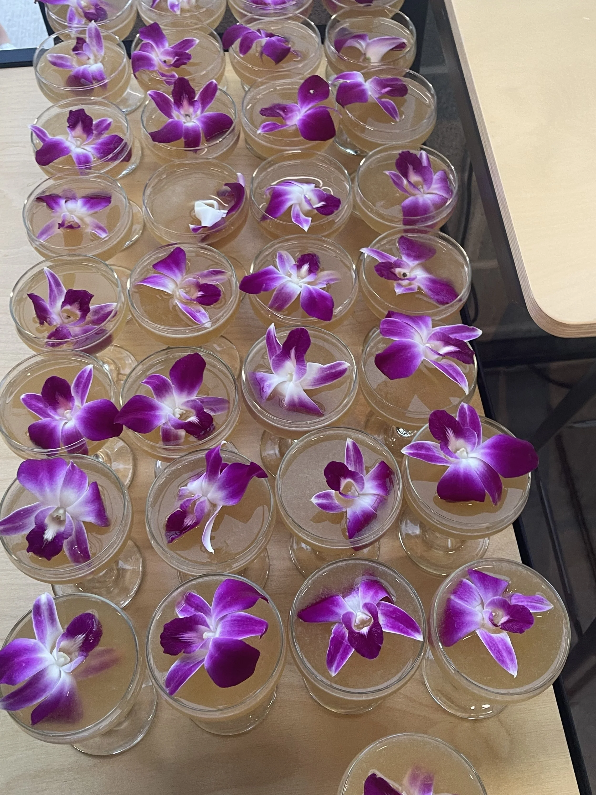 Multiple glasses of light-colored beverage topped with purple and white orchid flowers, arranged on a wooden surface.