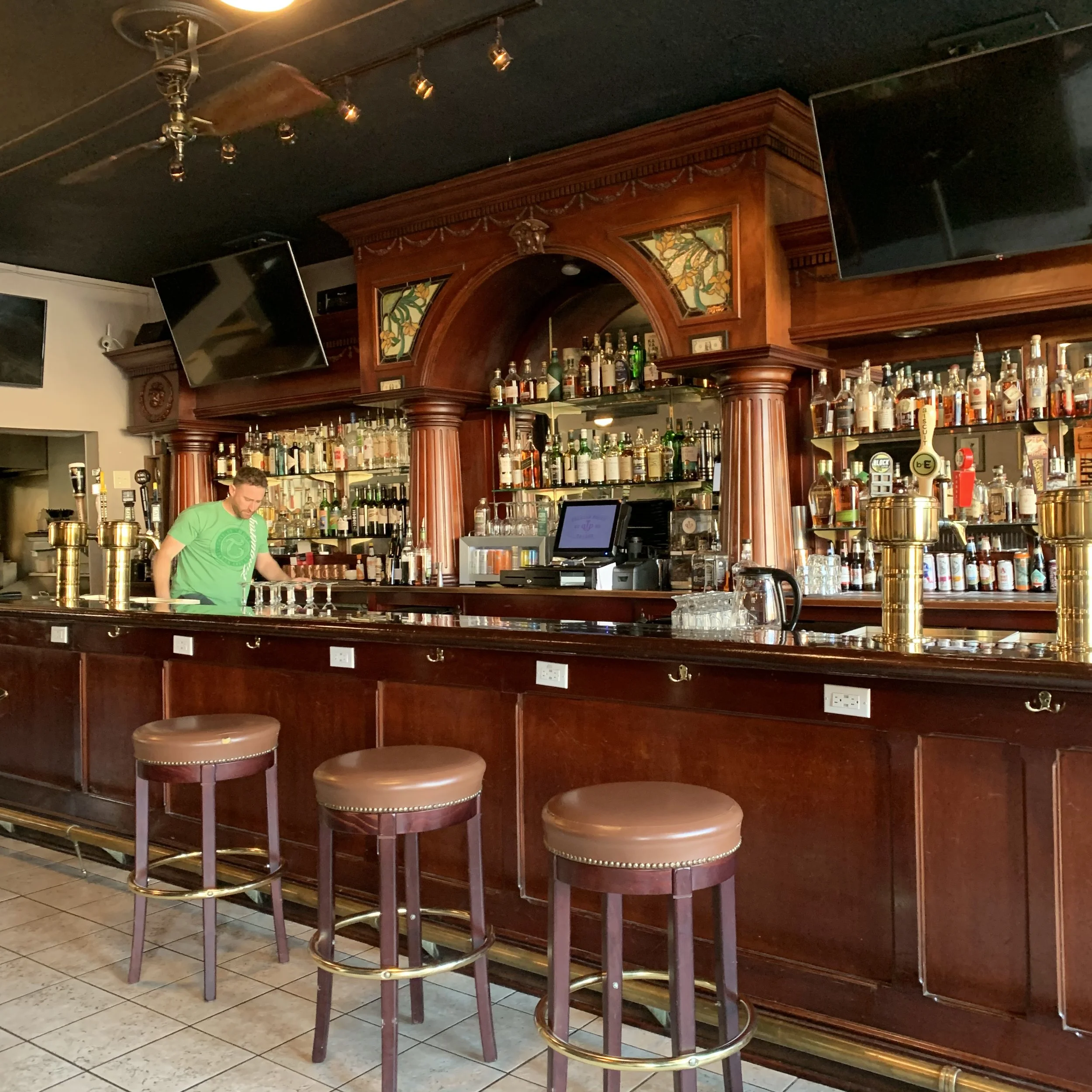 A bar with wooden counter, bar stools, multiple bottles of alcohol on shelves, and two television screens mounted on the wall.