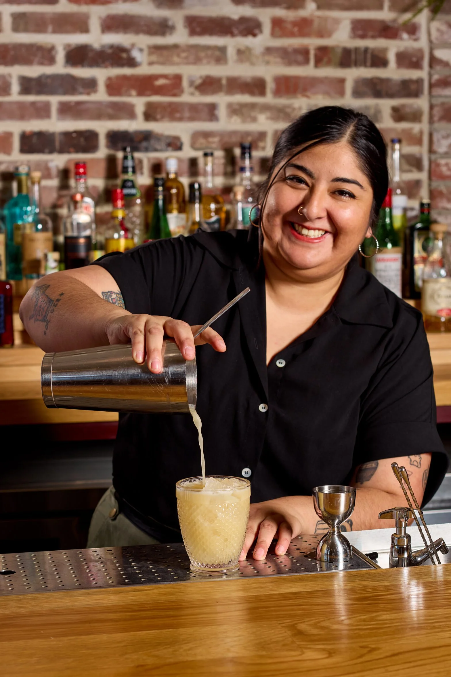 A smiling bartender with tattoos, wearing a black shirt, is pouring a drink into a textured yellow glass behind a bar with a brick wall and an assortment of liquor bottles in the background.