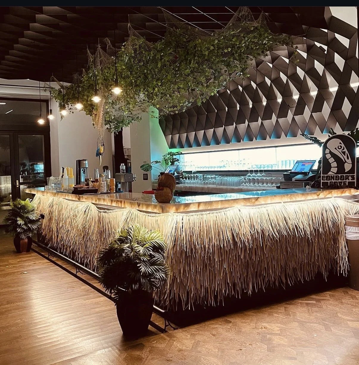 A tropical-themed bar with a straw skirted counter, potted plants, hanging greenery, and geometric wall panels, illuminated by warm lighting.