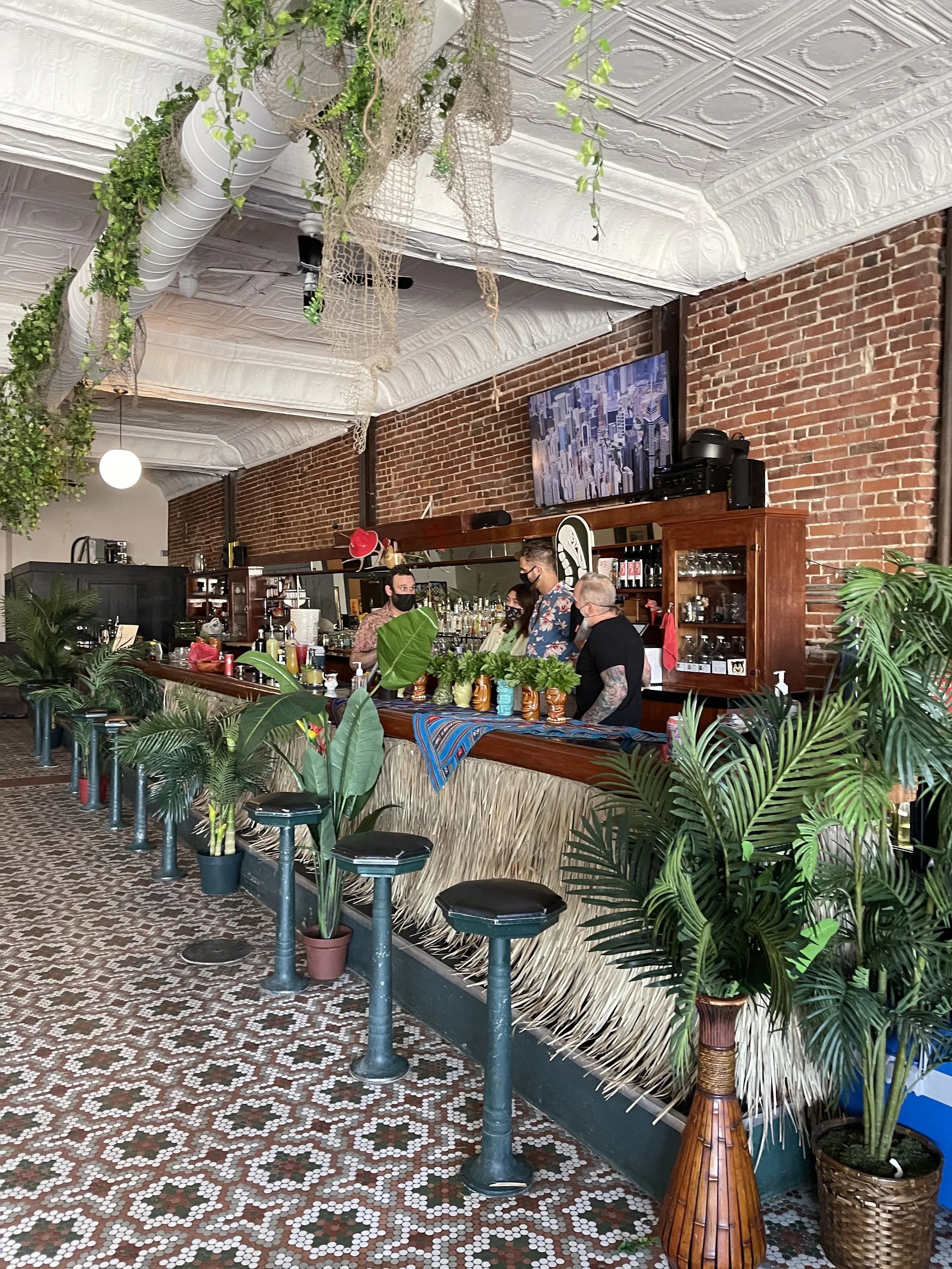 Inside a vibrant restaurant or bar with a vintage mosaic tile floor, a bar counter with plants and barware, brick walls, framed art, and patrons with masks, lush greenery and decorative elements.