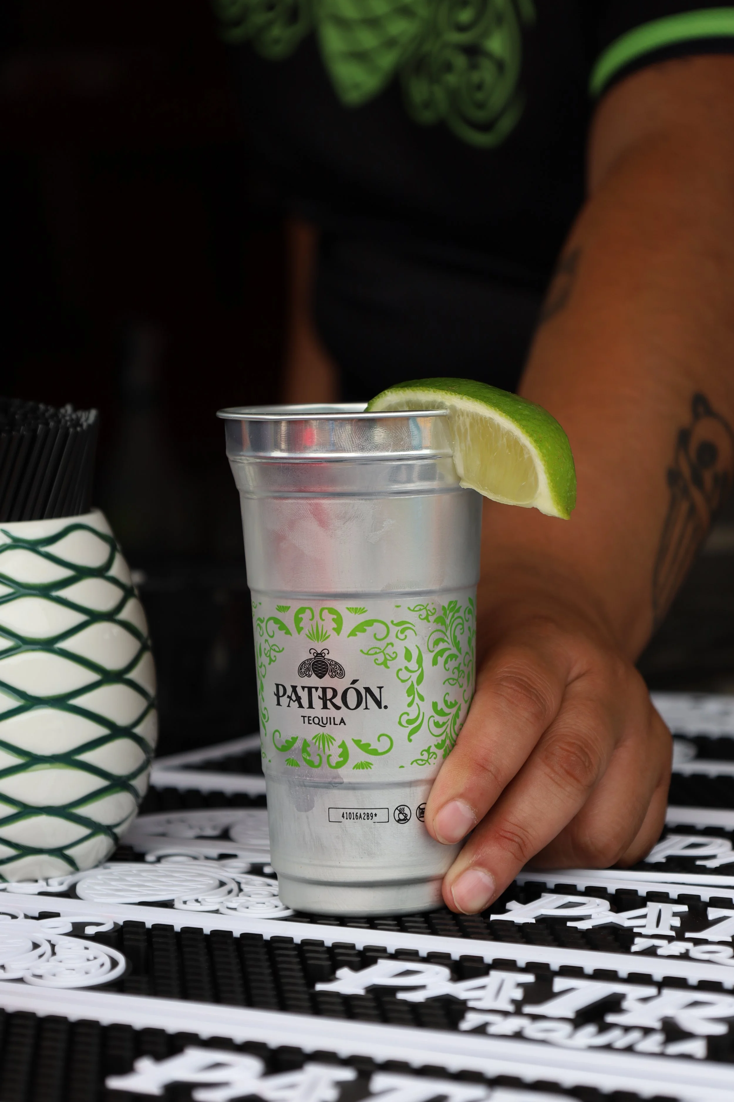 A hand holding a tall glass of clear liquid with a lime wedge garnishing the rim, with a Patrón tequila label visible on the glass.