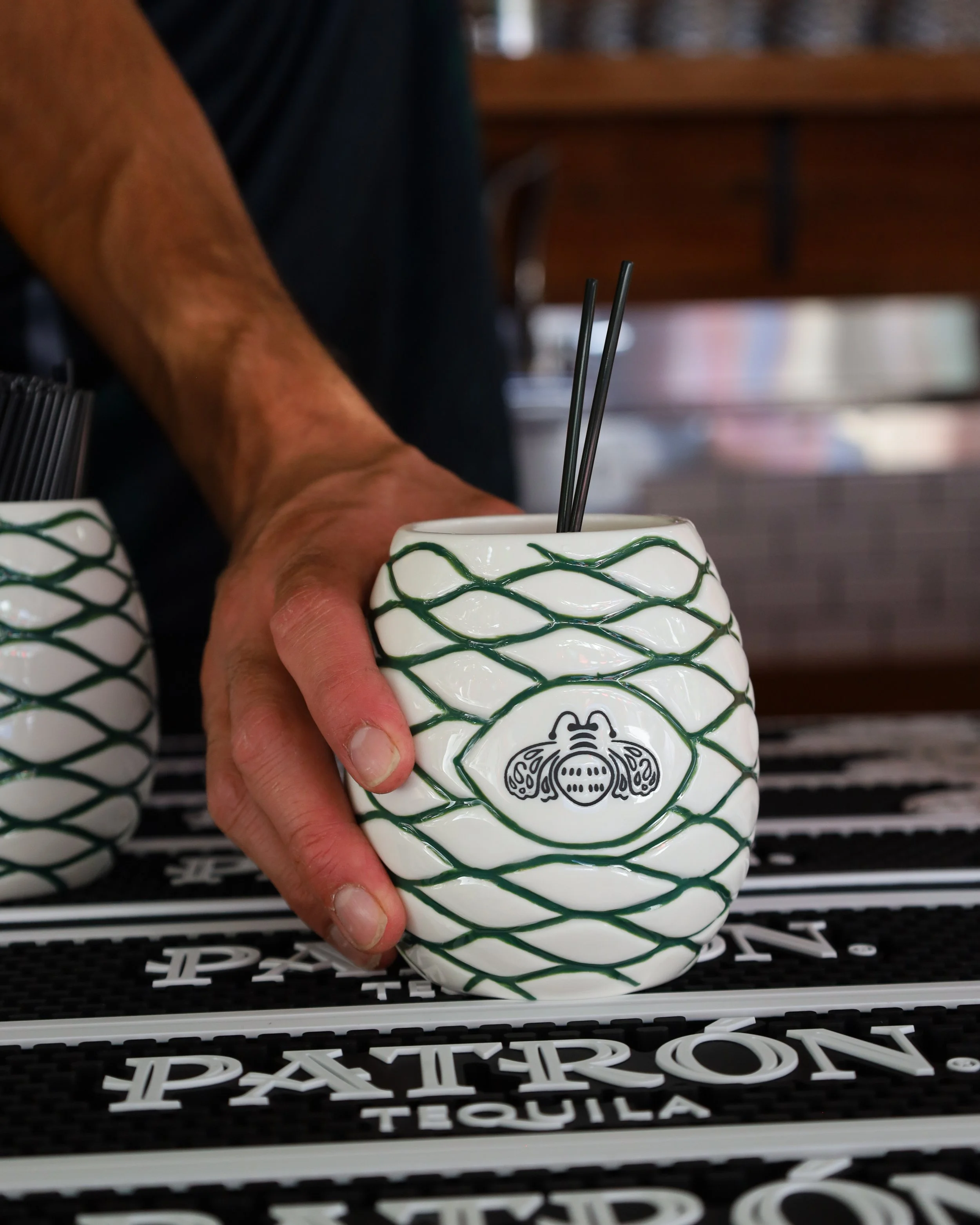 A person's hand holding a ceramic mug with a black and white tribal design, containing two black drinking straws, placed on a black surface with white text and patterns, with blurred background.