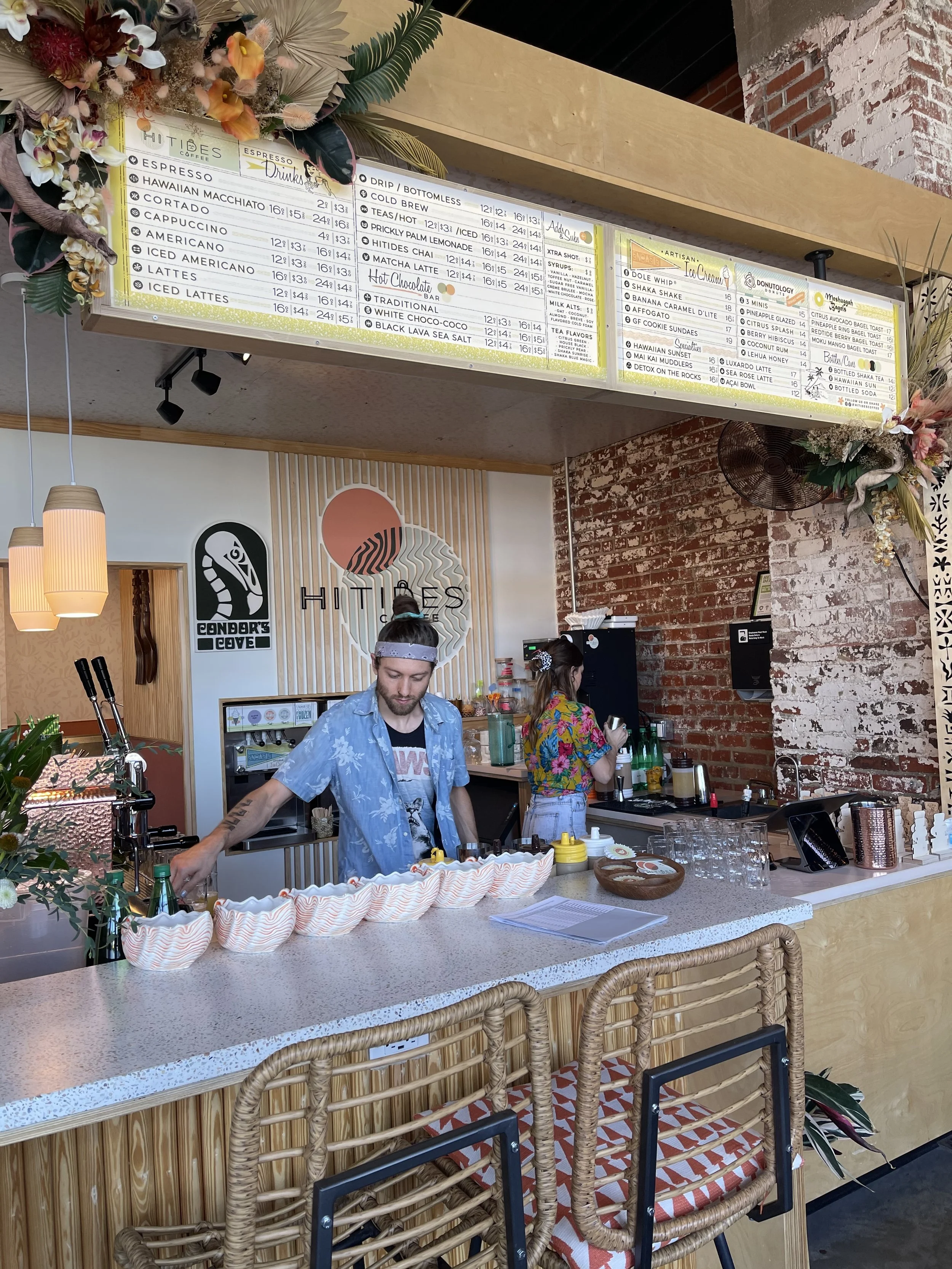 A cafe counter with two employees preparing drinks, decorated with flowers and tropical plants, with a menu board overhead displaying various coffee, tea, and specialty drinks.