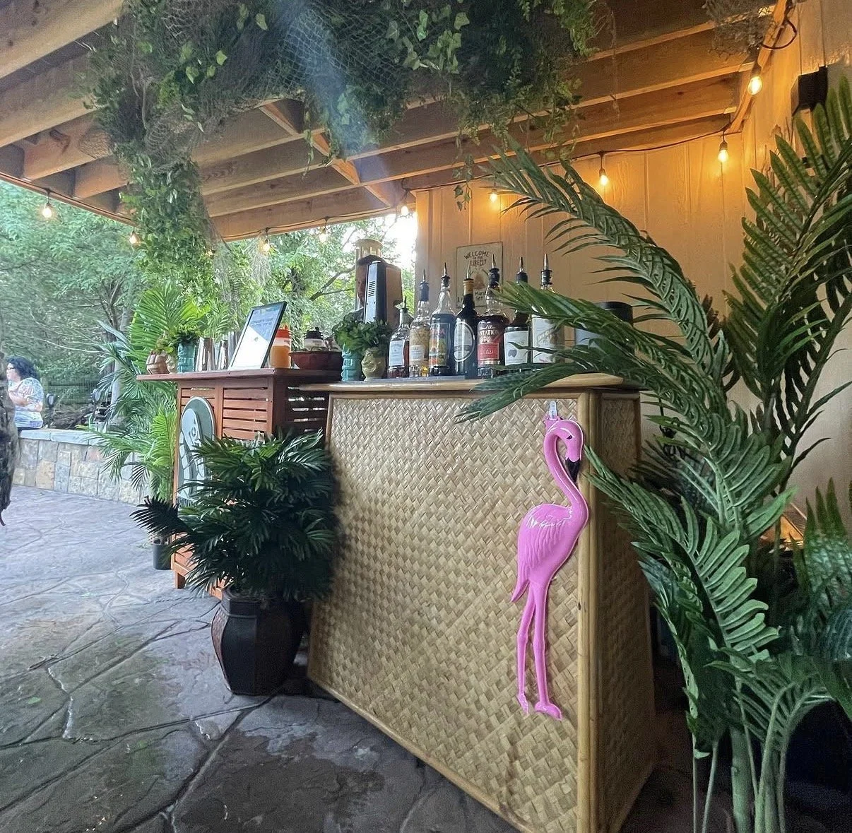 A tropical-themed bar area with a woven bamboo counter, surrounded by lush green plants, and decorated with a pink flamingo figure hanging on the side. The bar has various bottles and a small coffee machine, with warm string lights overhead and a sla