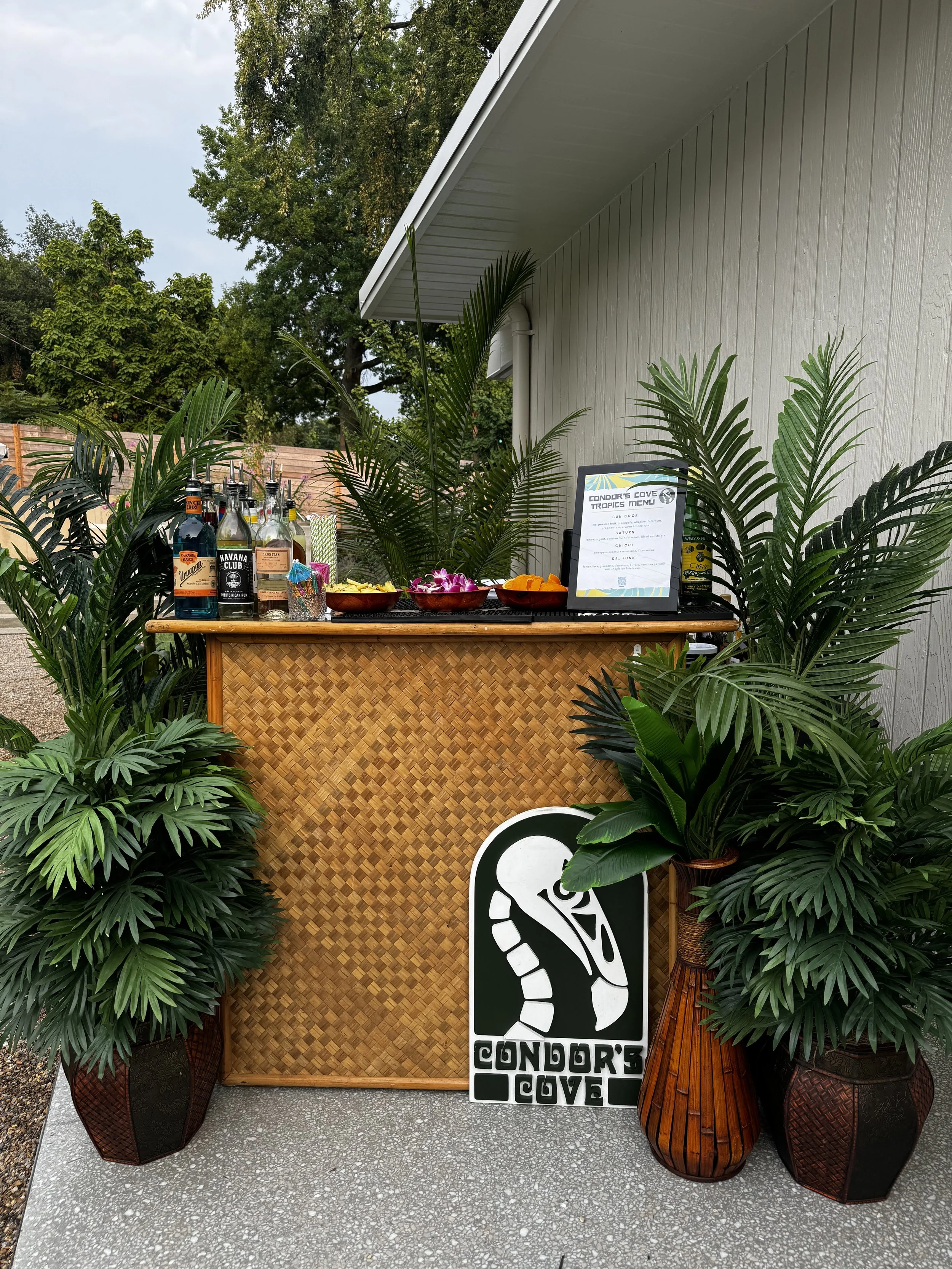 A tiki bar setup outdoors with a wooden woven front, tropical plants, bottles of alcohol on a small table, a sign with the text 'Condor's Cove' and a footprint logo, and a bamboo-style wooden table on the right.