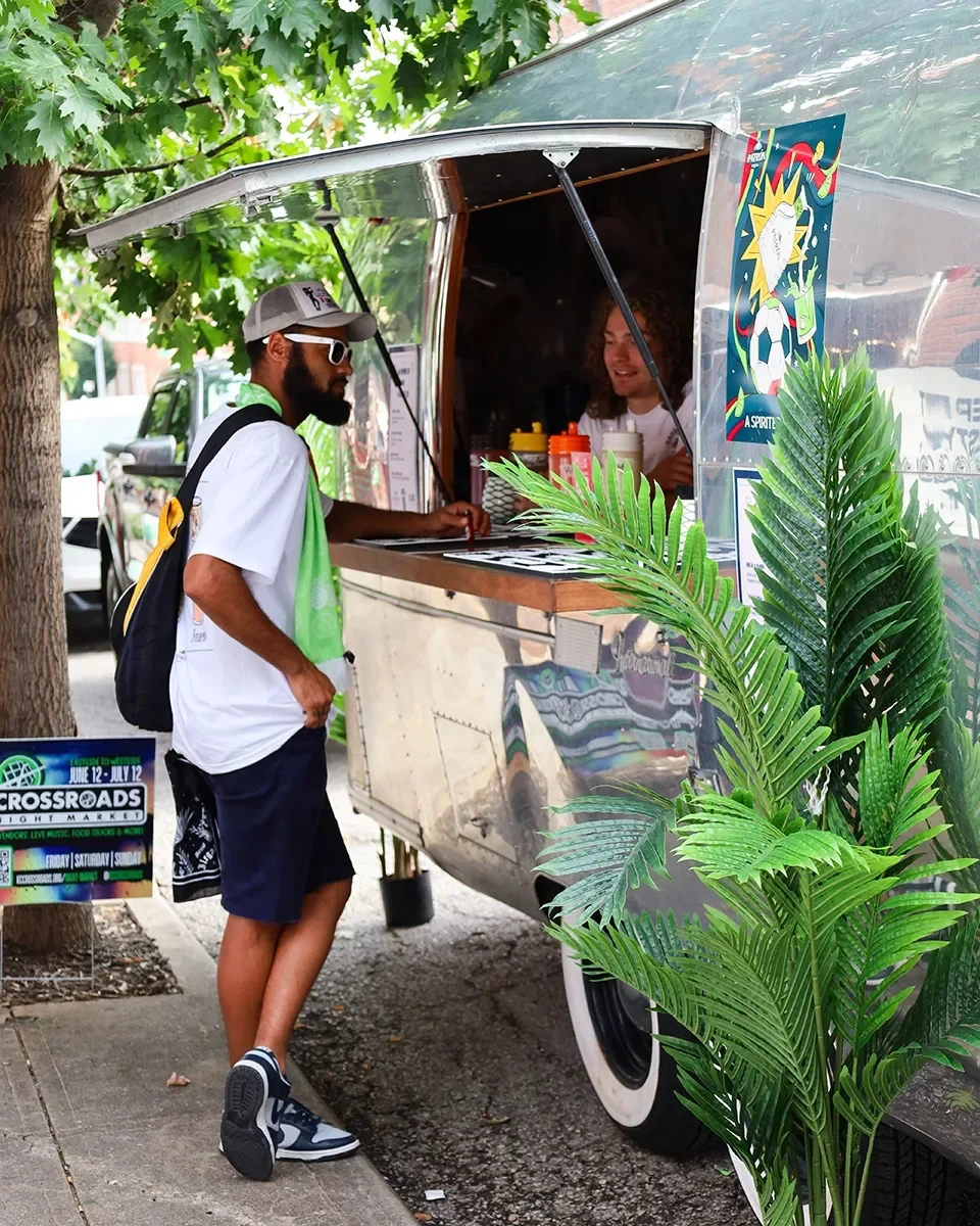 A man wearing a gray cap, white T-shirt, navy shorts, and sneakers orders food at a food truck. The food truck is decorated with colorful posters and plants, and a woman inside the truck helps him. There is a tree with green leaves and a sidewalk in 