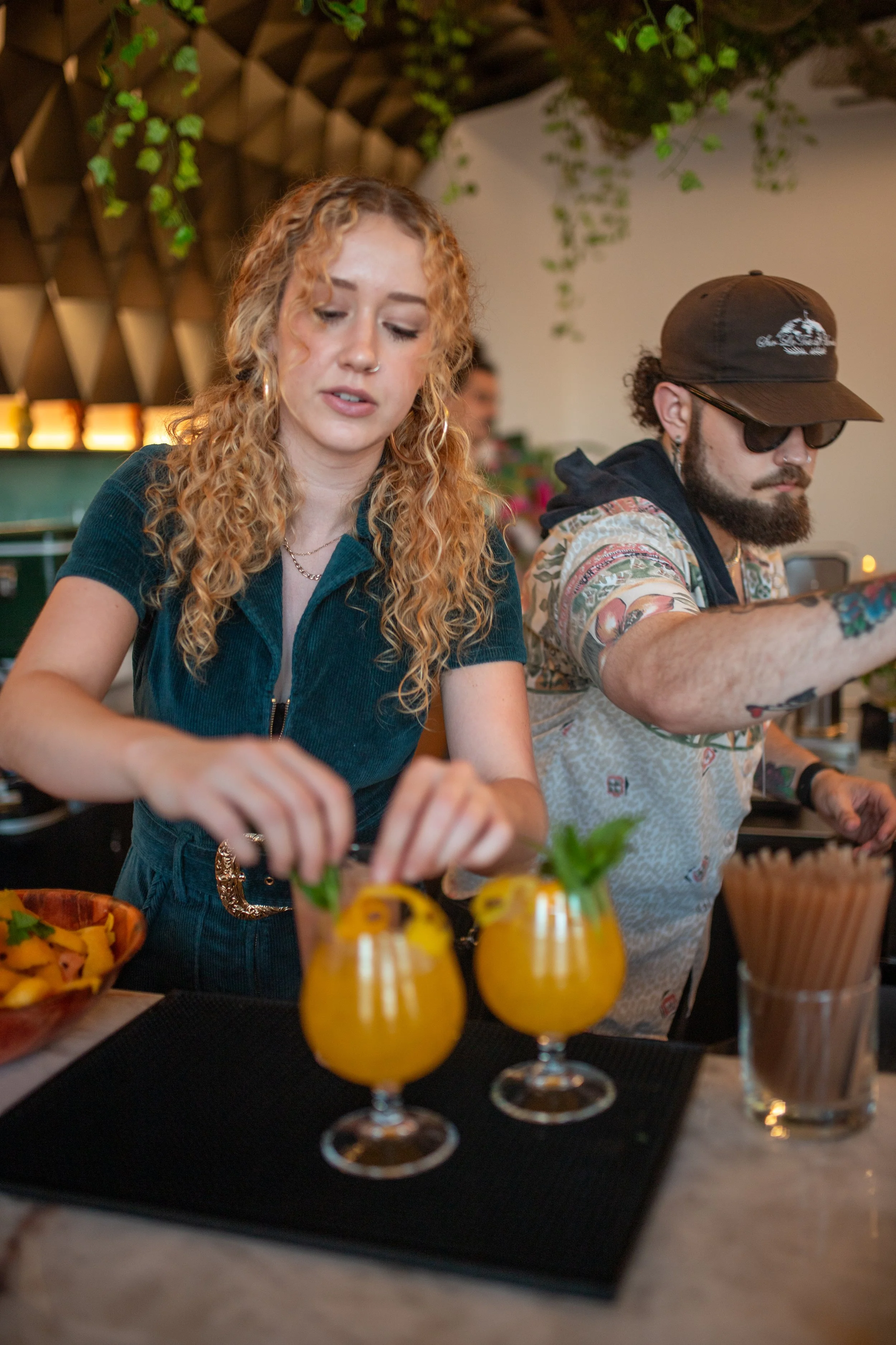 A woman with curly blonde hair, wearing a teal zip-up top, is preparing drinks at a table with two orange cocktails garnished with herbs. Beside her, a man with a beard, sunglasses, a brown cap, and a patterned shirt is reaching or working on somethi