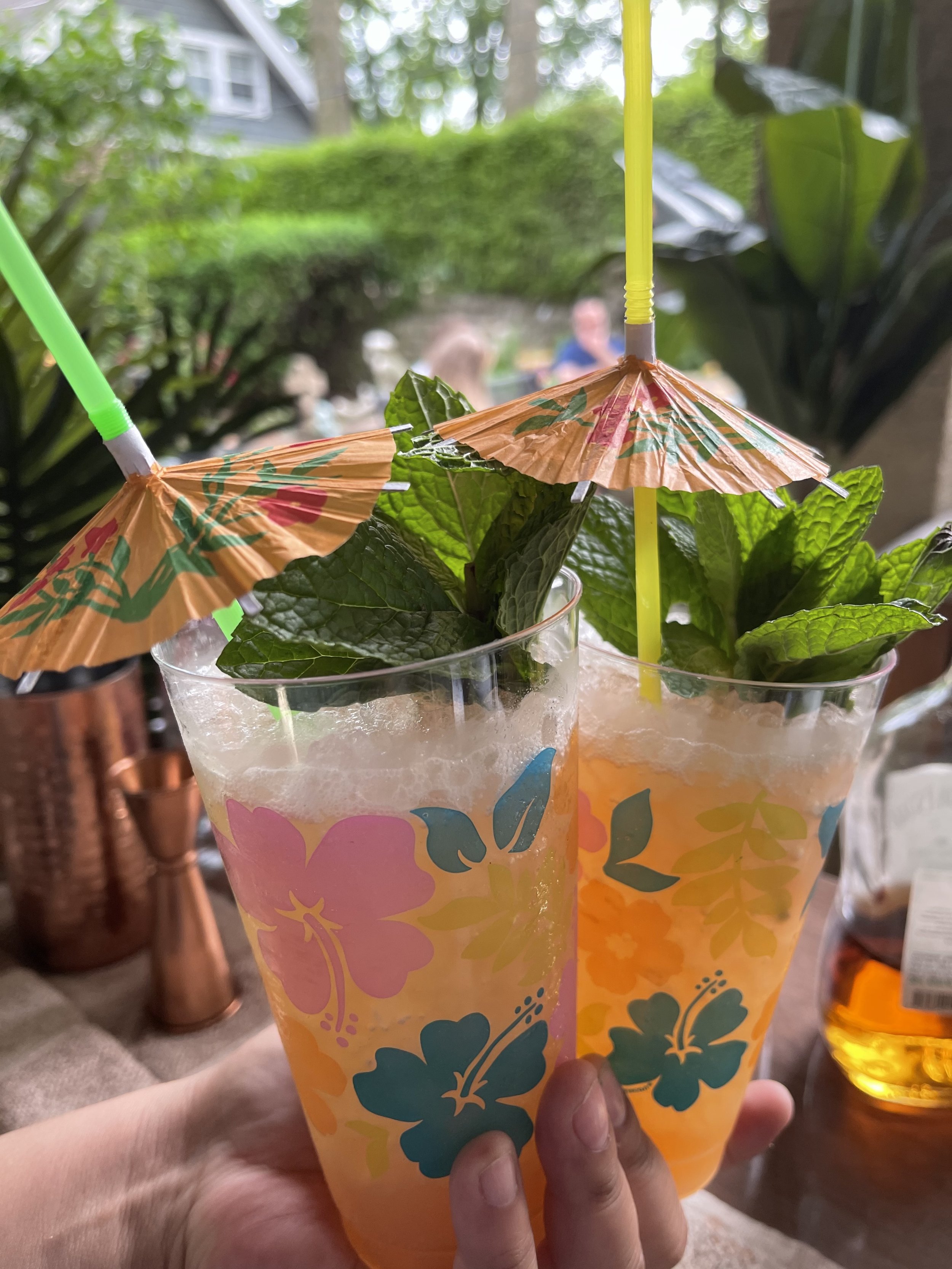 Two colorful cups with pink and orange floral designs, filled with a light-colored beverage and topped with crushed ice, are decorated with small paper umbrellas and straws. An outdoor background with greenery and a house is visible.