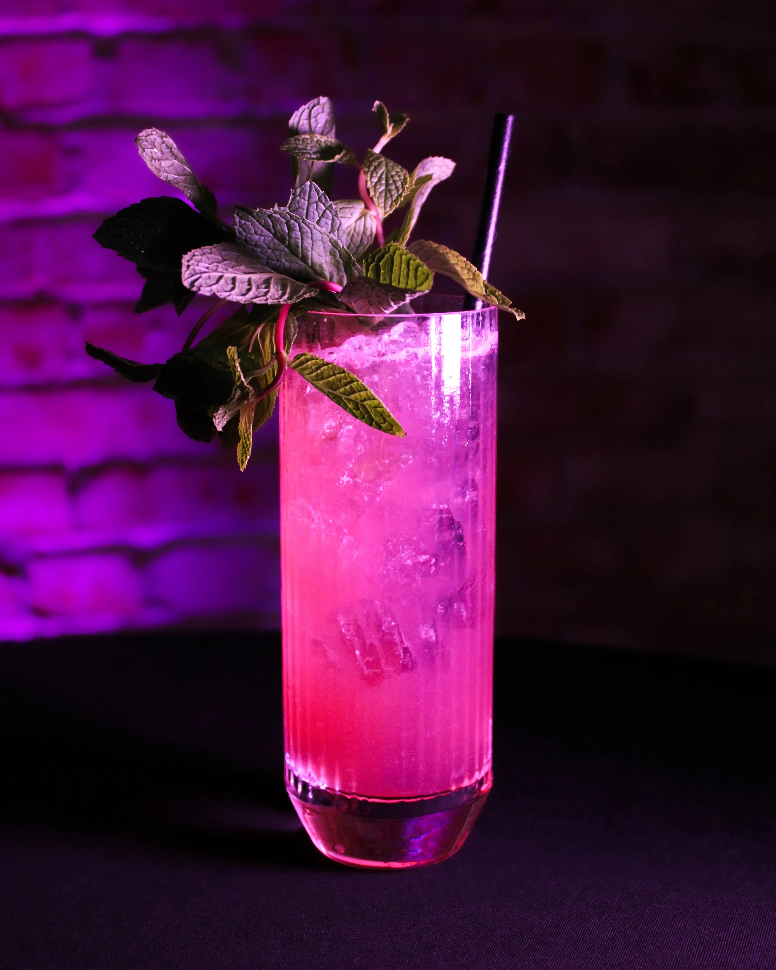 A pink glowing drink in a tall glass garnished with fresh mint leaves, with a black straw, against a dark purple background.