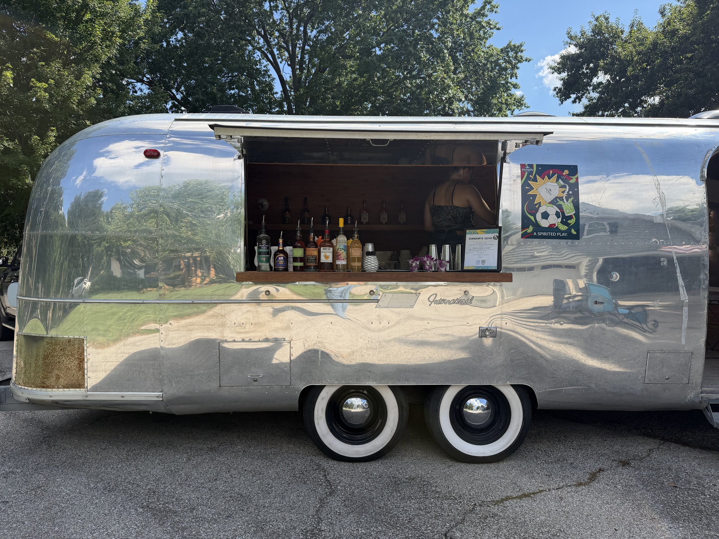A shiny, silver Airstream trailer with an open serving window. Inside, there are bottles of alcohol and cups on the counter. A woman in a strapless top is visible inside, preparing drinks. There are trees and a blue sky with some clouds in the backgr