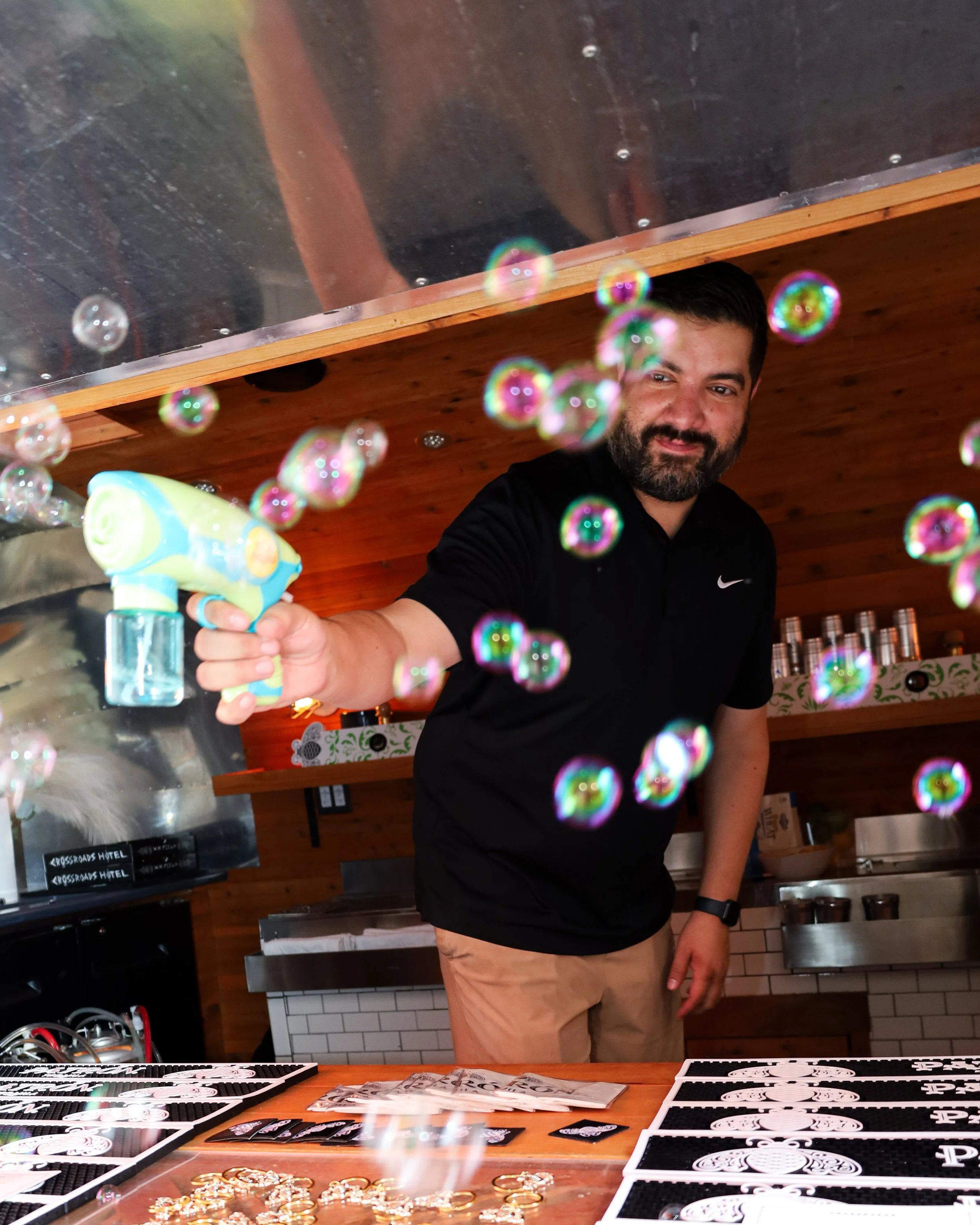 A man with dark hair and beard, wearing a black Nike t-shirt, is holding a bubble gun and spraying soap bubbles. He is inside a wooden-paneled room, with a display of jewelry and black and white cards on a table in front of him.