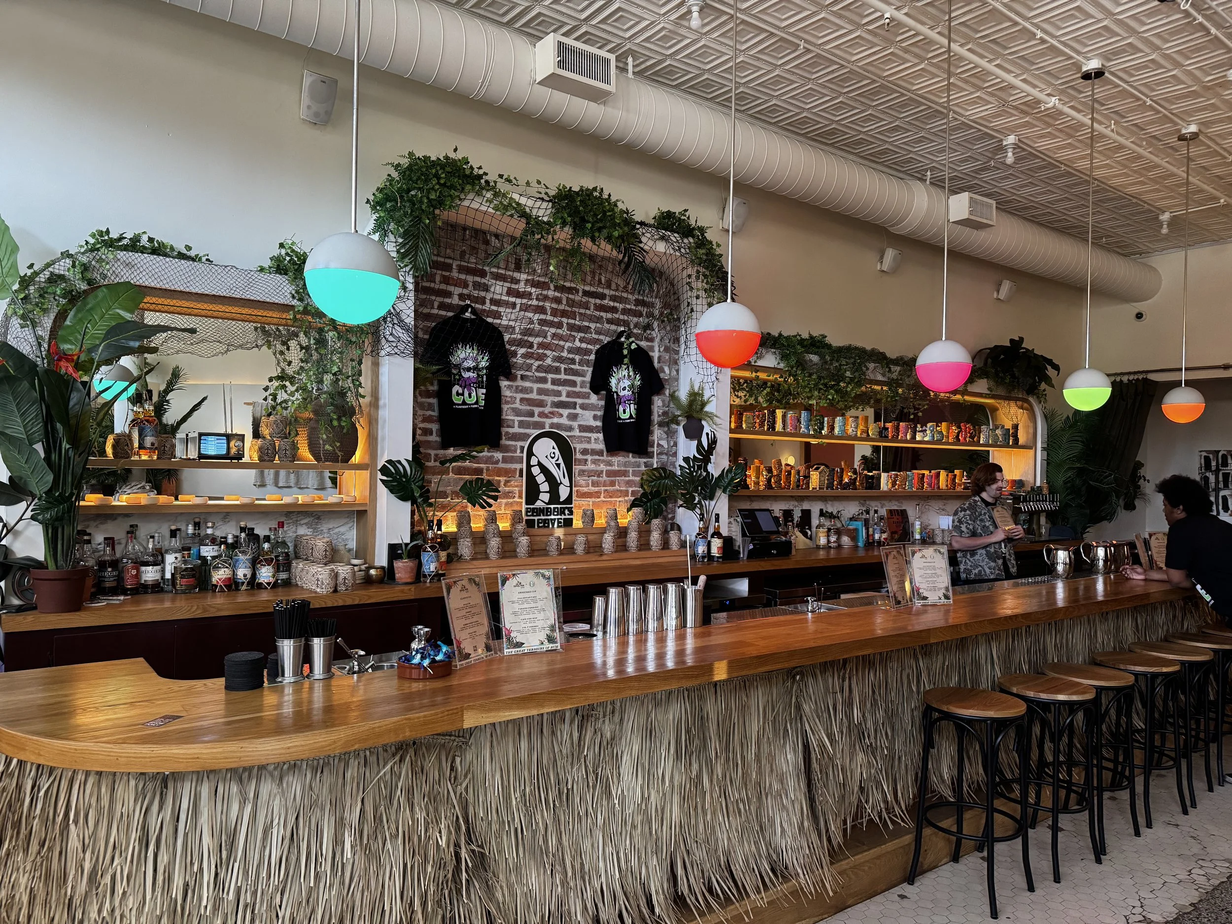 Interior of a bar or restaurant with a long wooden counter, barstools, tropical plants, hanging colorful spherical lights, and a brick wall background with T-shirts and shelves of colorful bottles.