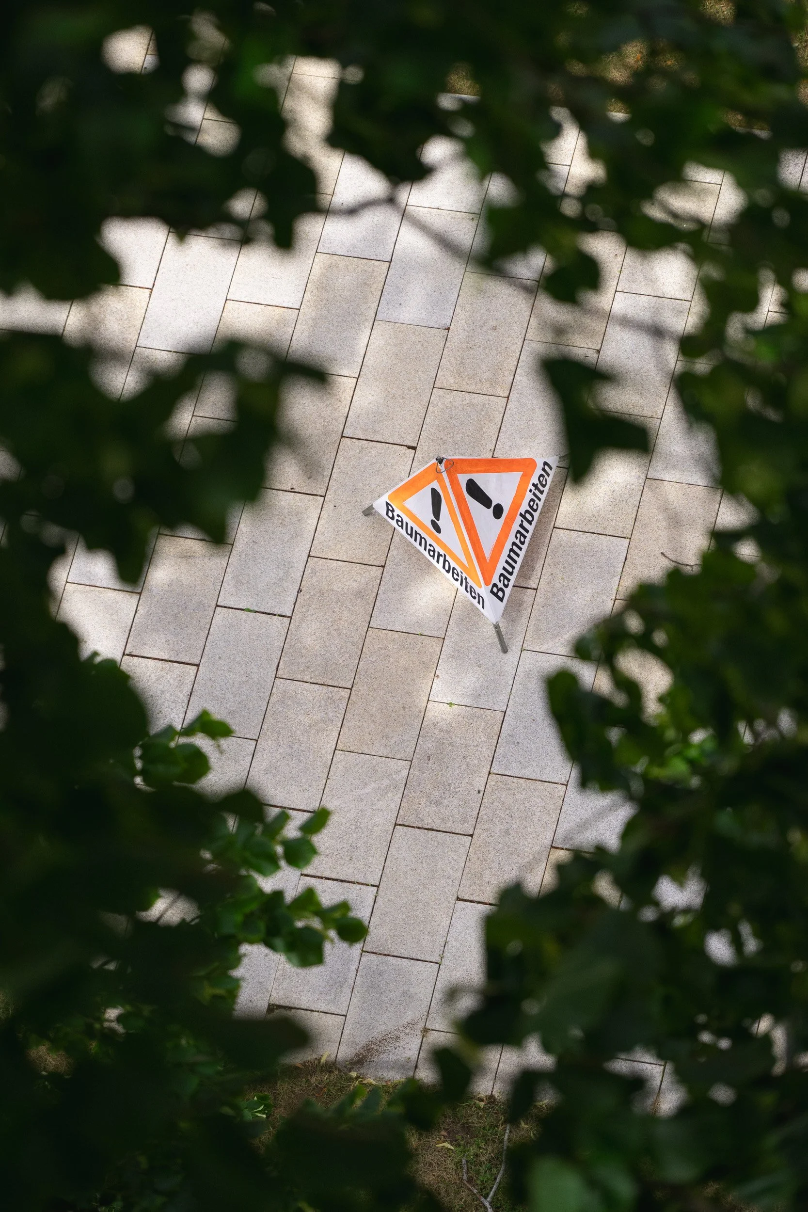 Warnschild „Baumarbeiten“ liegt auf einem gepflasterten Weg, umgeben von grünen Blättern.