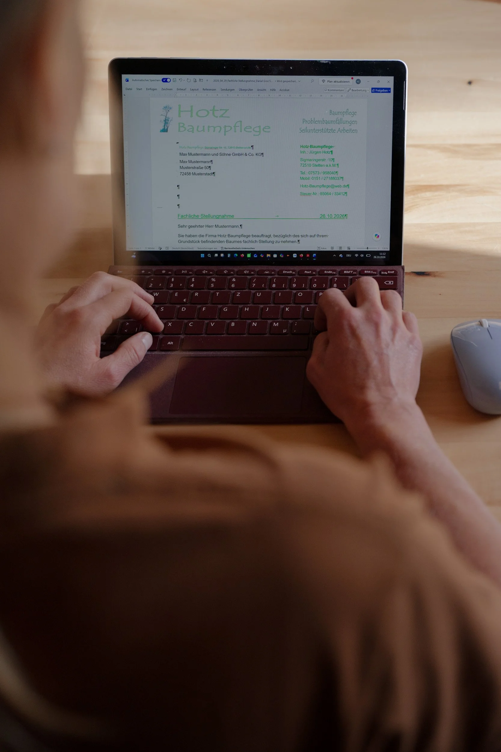 Person tippt an einem Laptop auf einem Holztisch an einem deutschen Word-Dokument über Baumpflege.