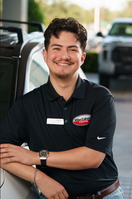 A smiling young male car dealership employee in a black polo shirt with a Toyota logo and name tag, standing outdoors in front of cars at a dealership.