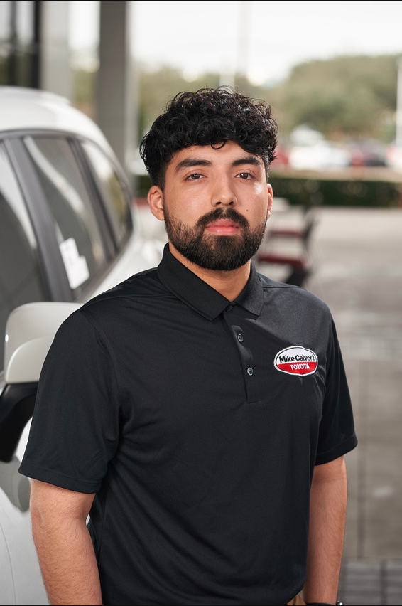 A man with curly dark hair and a beard wearing a black polo shirt with a Toyota logo standing outdoors near a white vehicle, with a parking lot and trees in the background.
