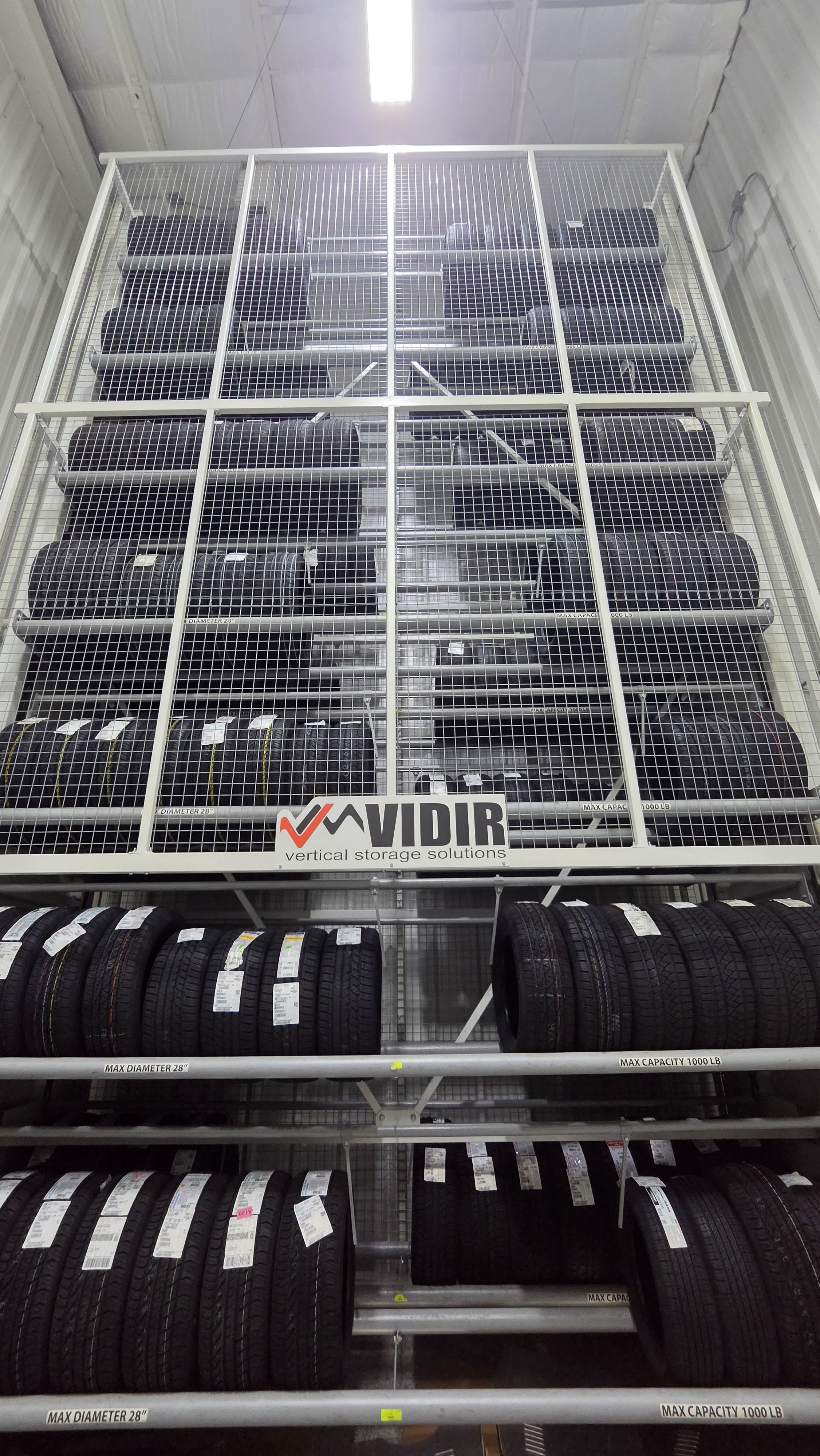 Tall metal shelving unit with four levels of tires stored on it, surrounded by a wire mesh in an industrial setting. The brand name 'Vidir' is visible on the front of the unit, along with the phrase 'vertical storage solutions'.