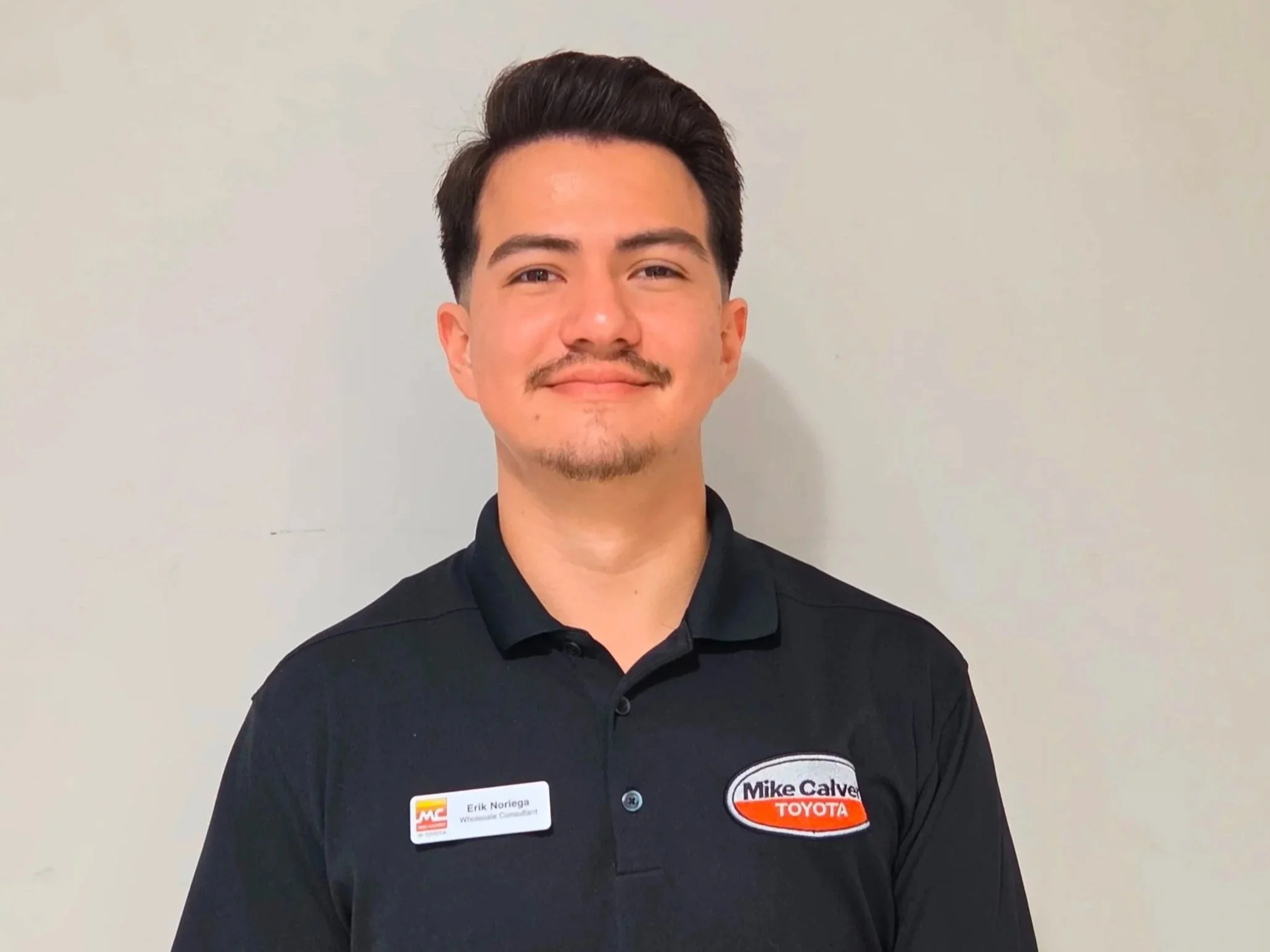 A young man with dark hair and a mustache wearing a black polo shirt with a name tag and a Toyota sponsorship patch, standing in front of a plain beige wall.