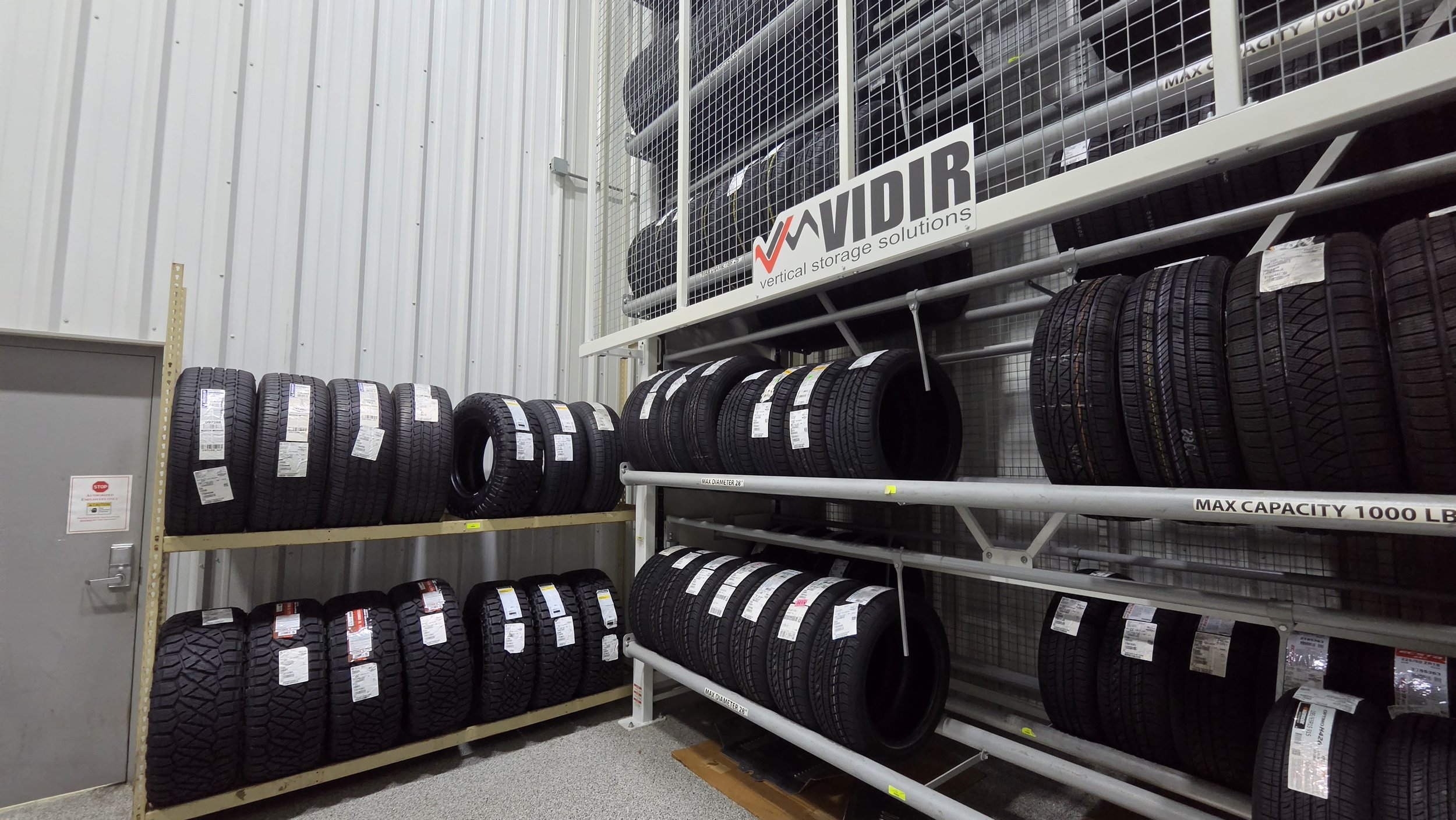 Shelf stocked with various car tires in a storage room, with a sign reading 'Vivid vertical storage solutions' and labels on the tires.