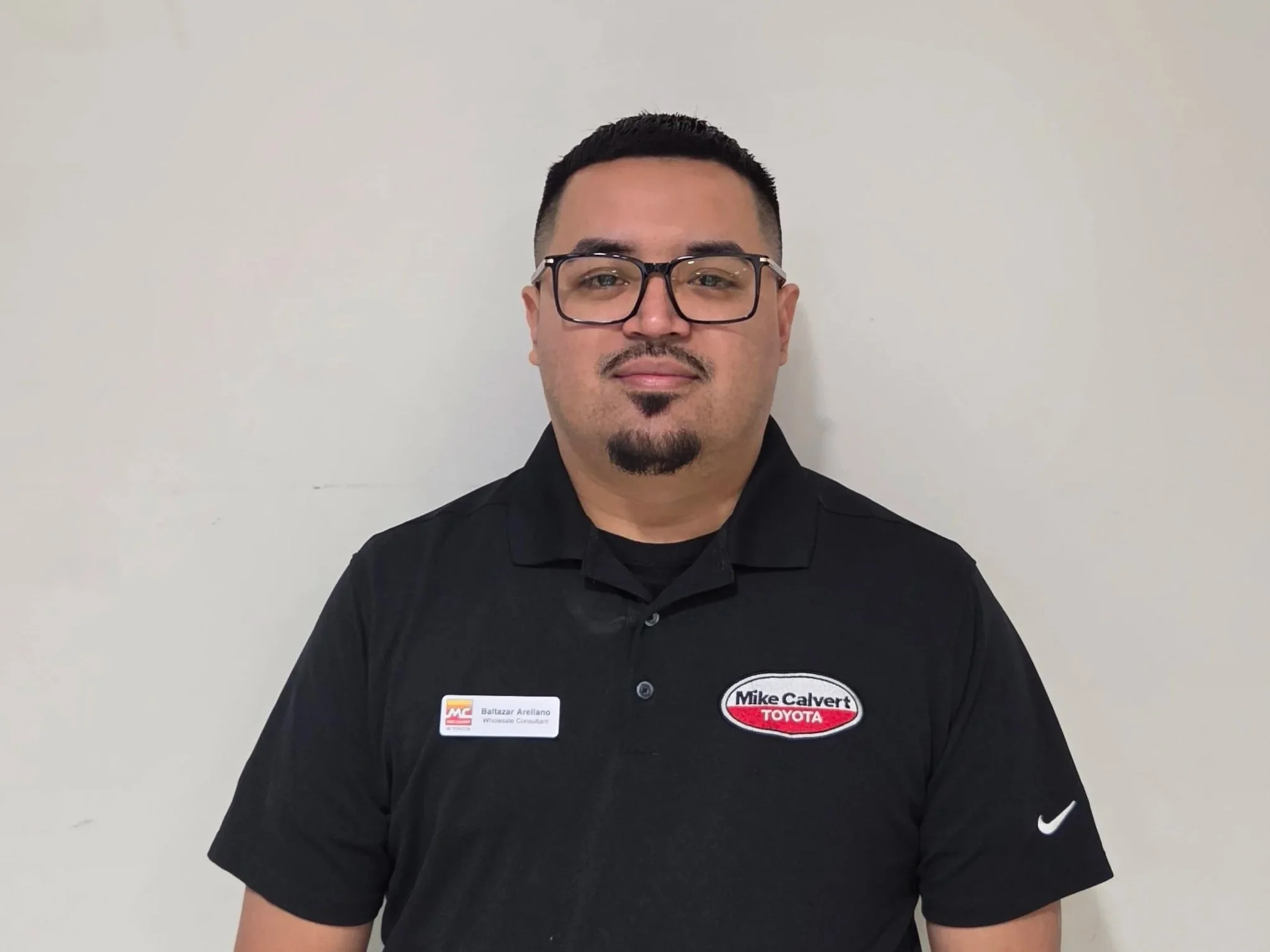 A man with glasses, black hair, and a goatee, wearing a black shirt with a name tag and a logo badge for Mike Calvert Toyota, standing against a plain white wall.
