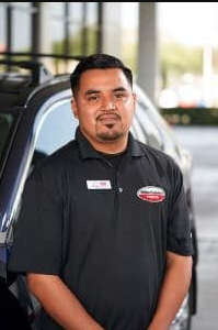 A man in a black uniform with a name tag, standing in front of a car.