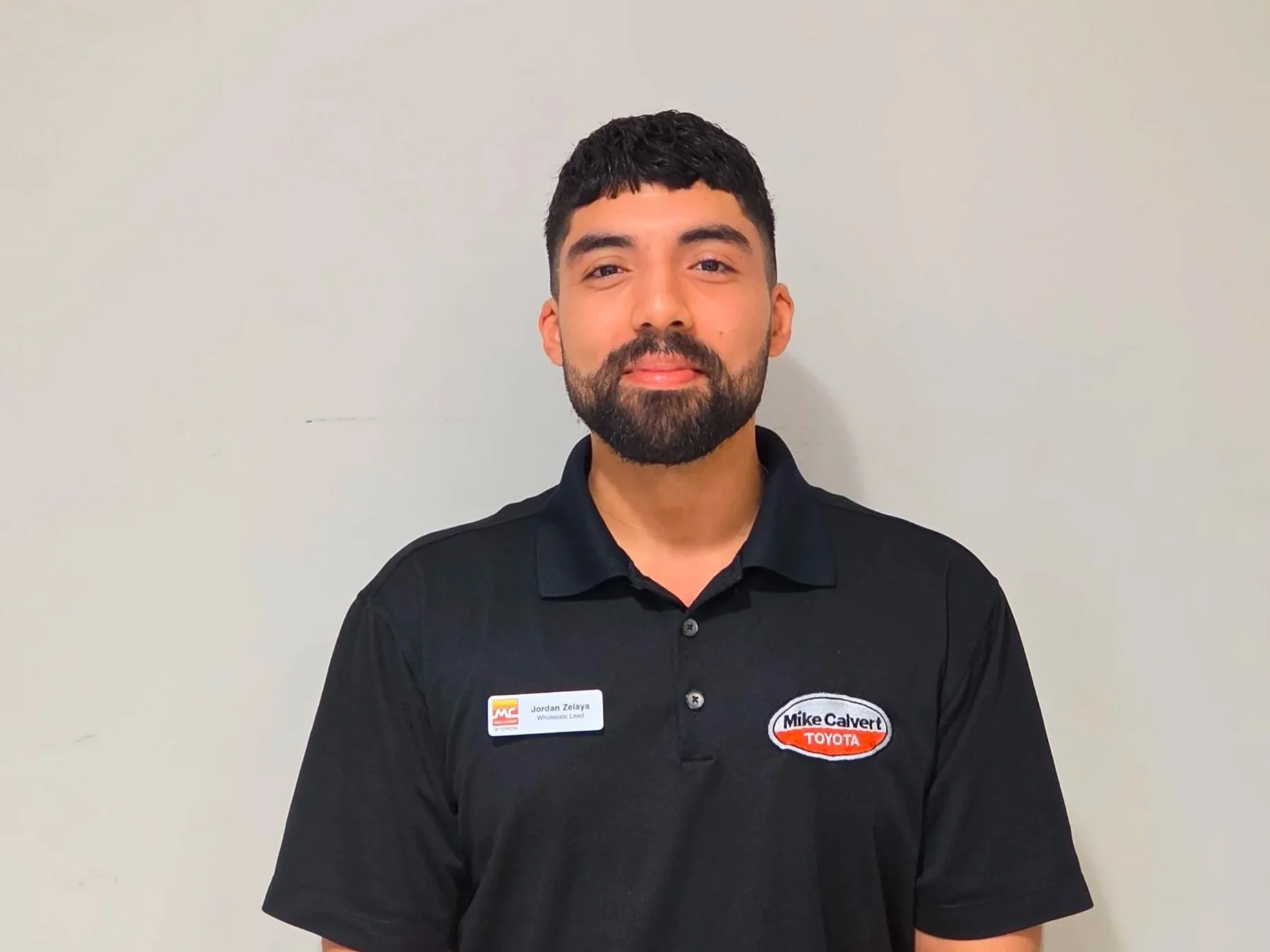 A man with a beard and short dark hair wearing a black collared shirt with a name tag and a circular patch, standing against a plain white wall.