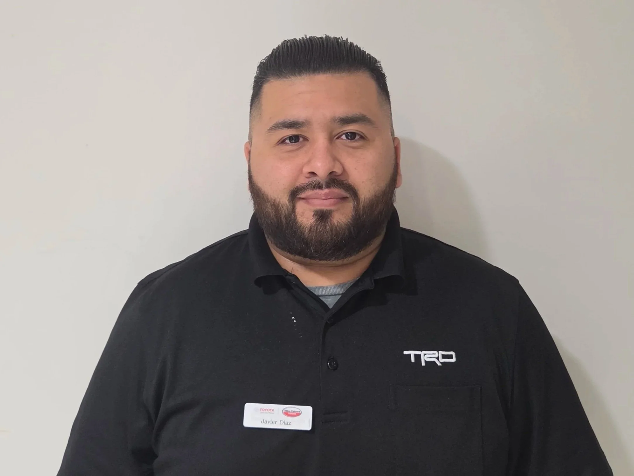 A man with a beard and short dark hair, wearing a black shirt with a Toyota and TRD logo, standing against a plain light-colored wall.