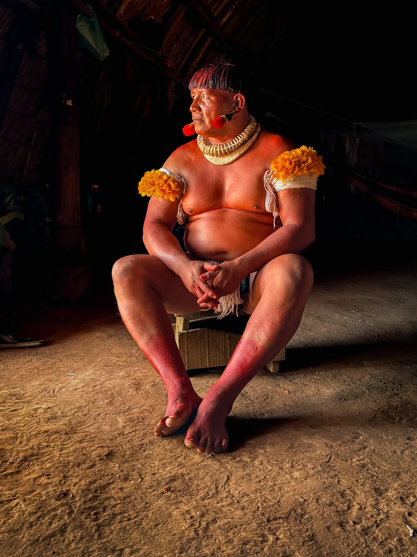 Un homme indigène vêtue de garde de fleurs jaunes sur les épaules et de colliers en coquillages, assis en méditation ou contemplation dans un espace intérieur sombre.
