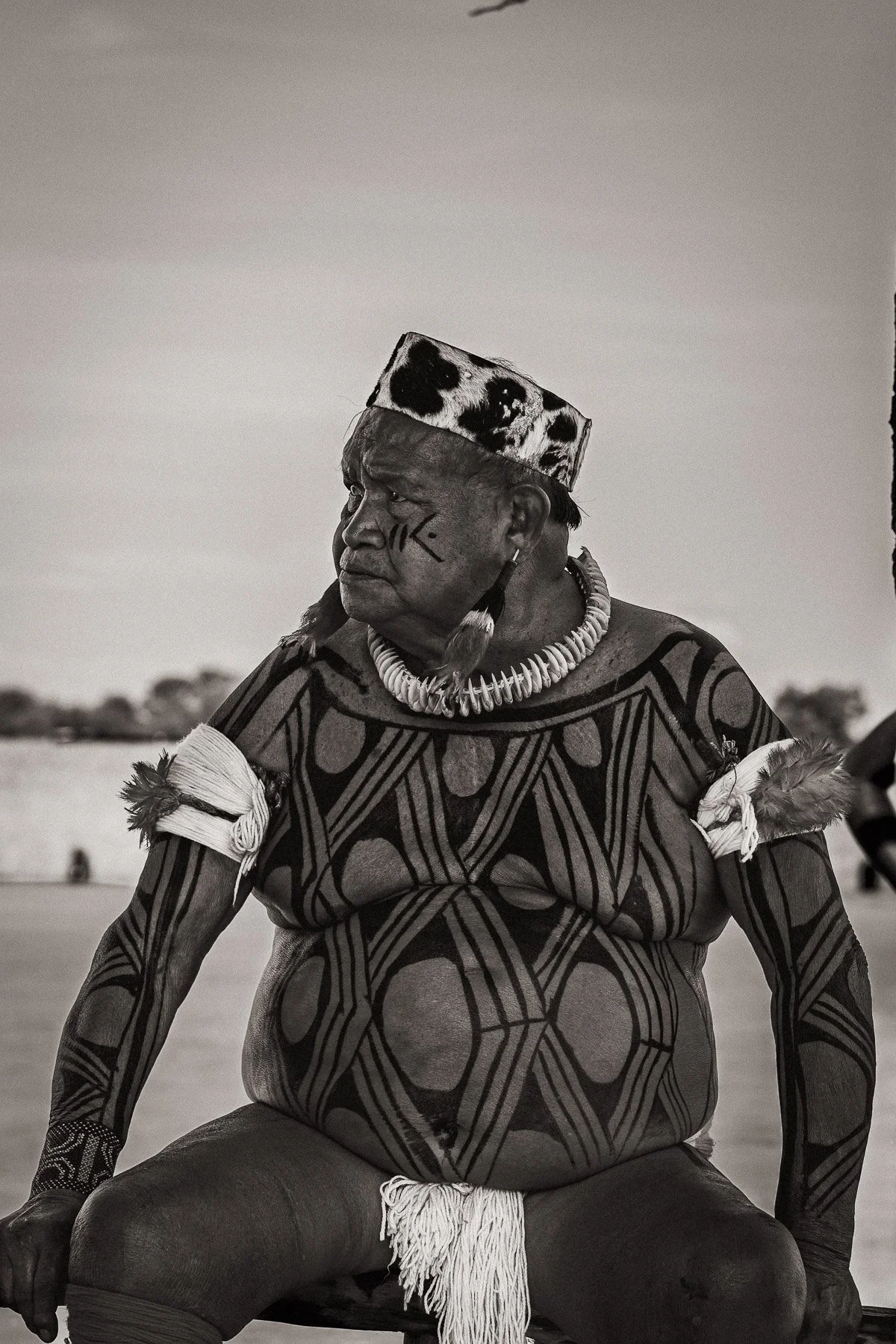 Un homme autochtone avec des tatouages traditionnels, portant un collier de coquillages et un chapeau à motif animal, assis dans un paysage désertique.