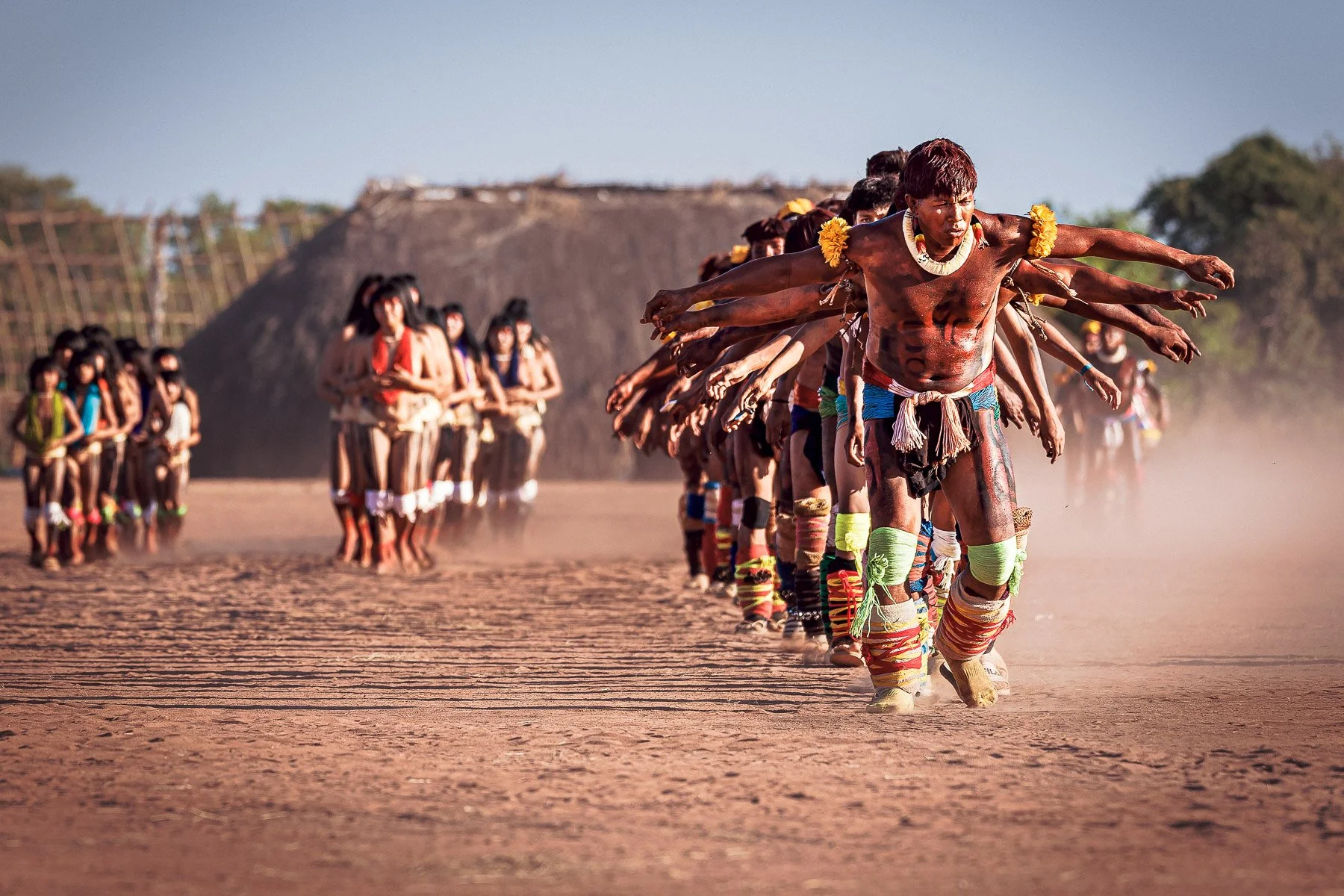 Groupe d'hommes et de femmes indigènes marchant en rang sur un terrain poussiéreux, portant des vêtements traditionnels et des accessoires colorés, avec une maison en terre en arrière-plan.