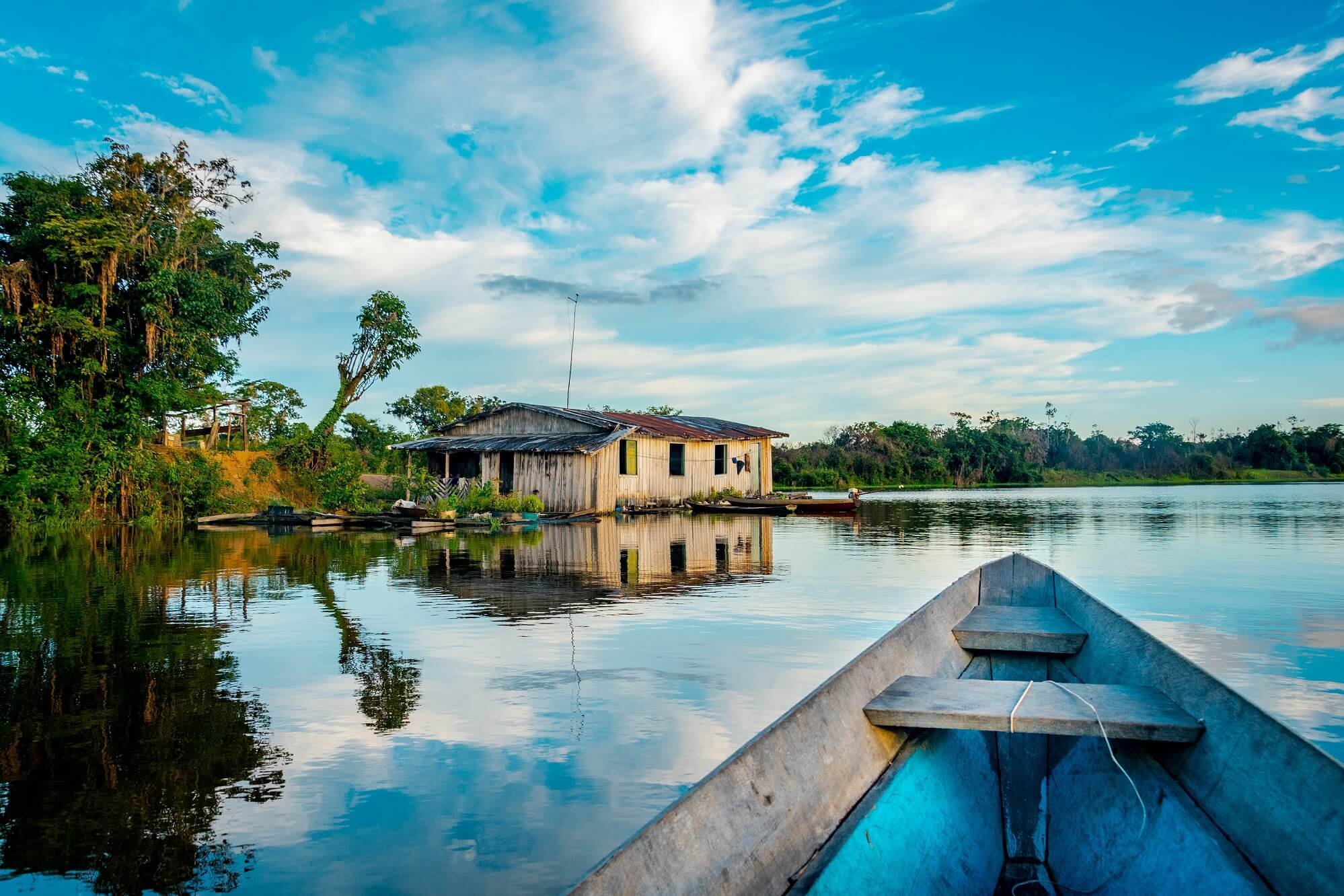 Une maison en bois au bord d'une rivière, entourée d'arbres, avec un ciel bleu et des nuages. Partiellement vue d'un bateau en bois dans l'eau.