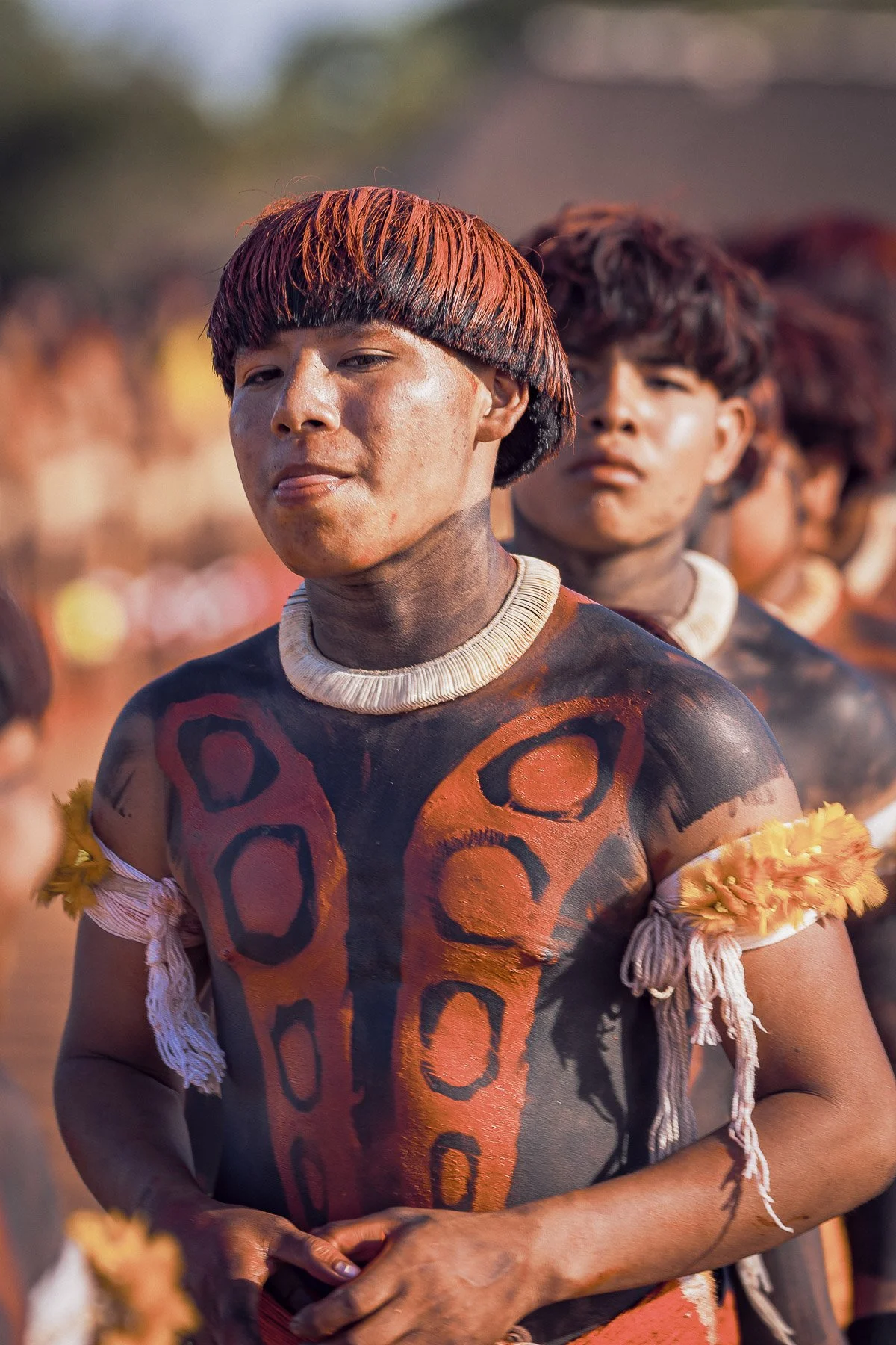 Jeune homme tatoué avec de la peinture orange et noire sur le corps, portant un collier en coquillage, lors d'une cérémonie ou fête sur fond flou.