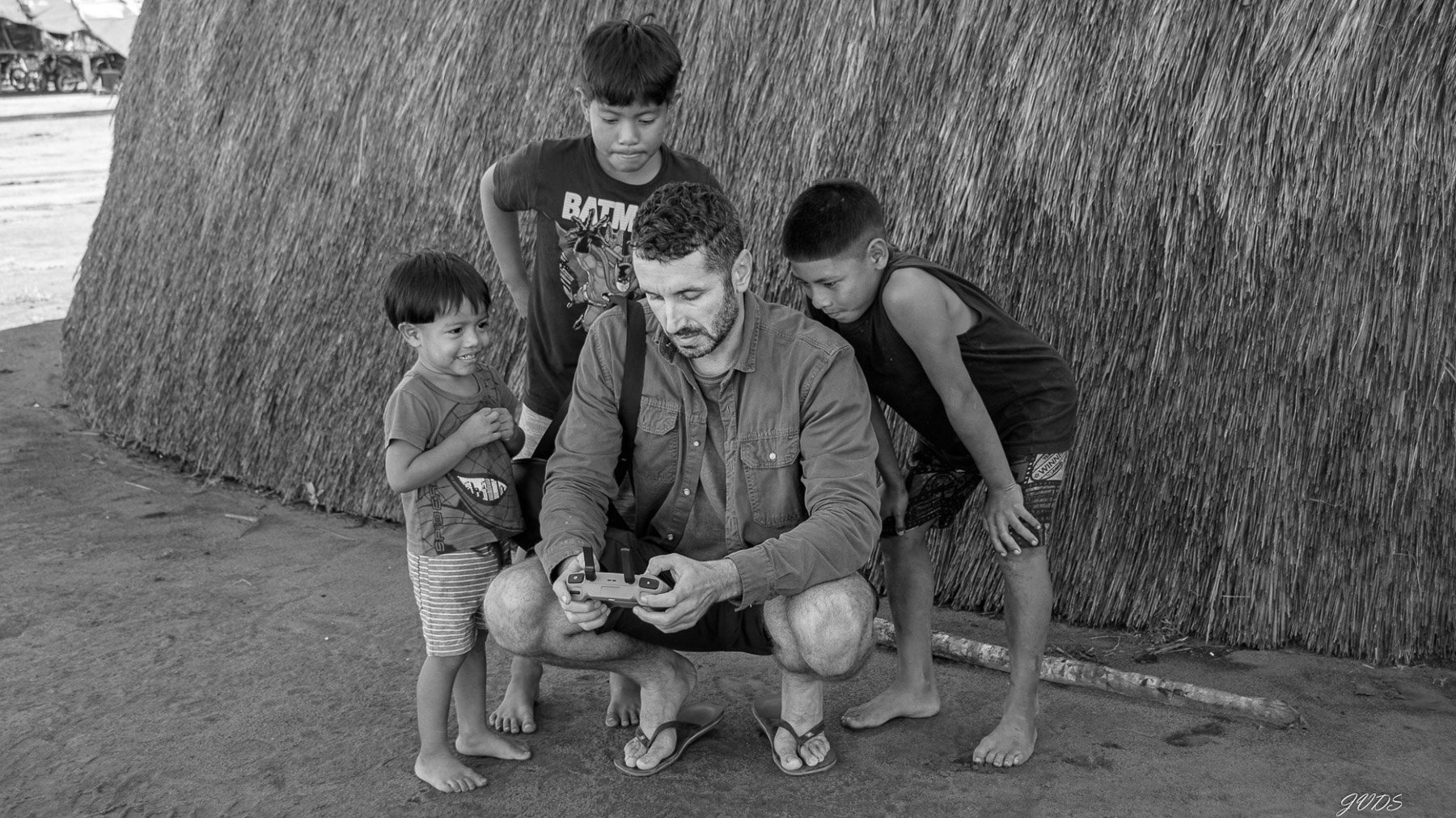 Un homme assis sur le sol, tenant une manette de jeu vidéo, entouré de quatre enfants, devant un mur en paille. La scène est en noir et blanc.