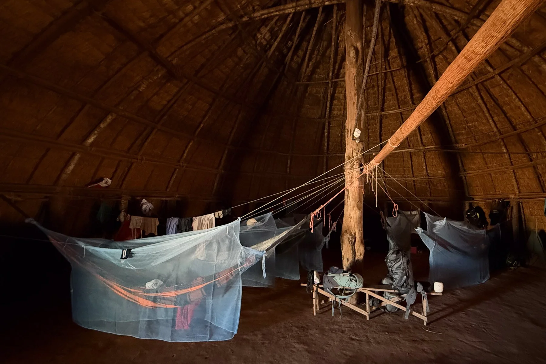 Intérieur d'une grande structure en bois avec un plafond en forme de dôme. Des draps ou toiles légères sont suspendus, intercalés avec des vêtements. On distingue également des sacs à dos et autres objets au sol.