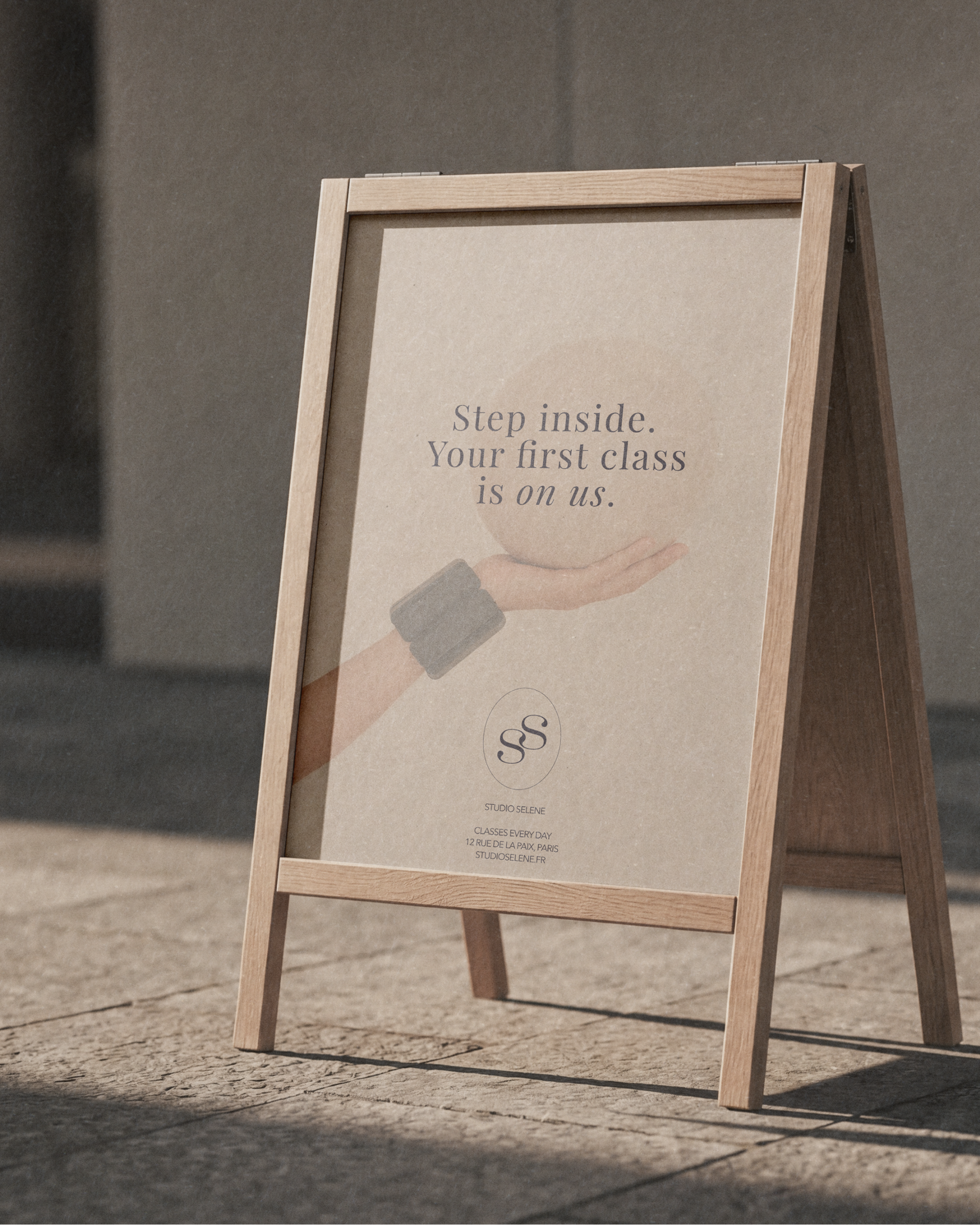 A wooden A-frame signboard with a beige poster that reads 'Step inside. Your first class is on us.' in dark gray text, standing outside on a concrete surface.