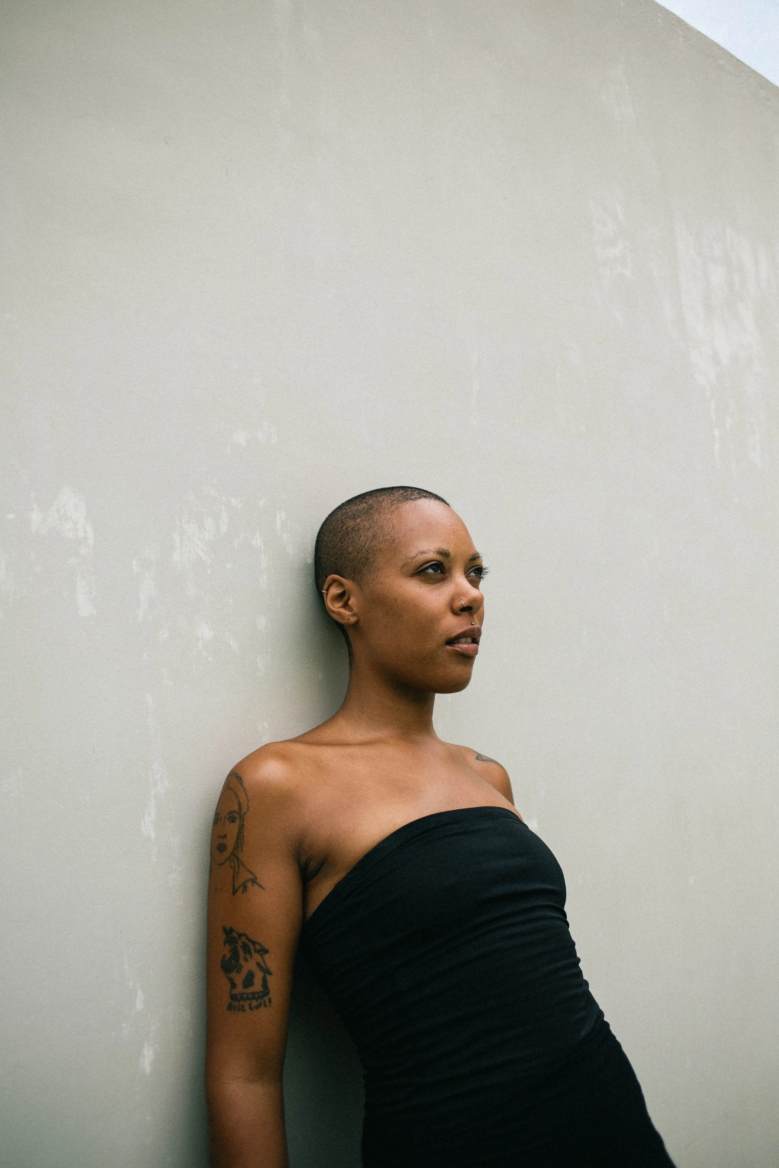 A woman with a shaved head and tattoos standing against a plain wall, wearing a strapless black top.
