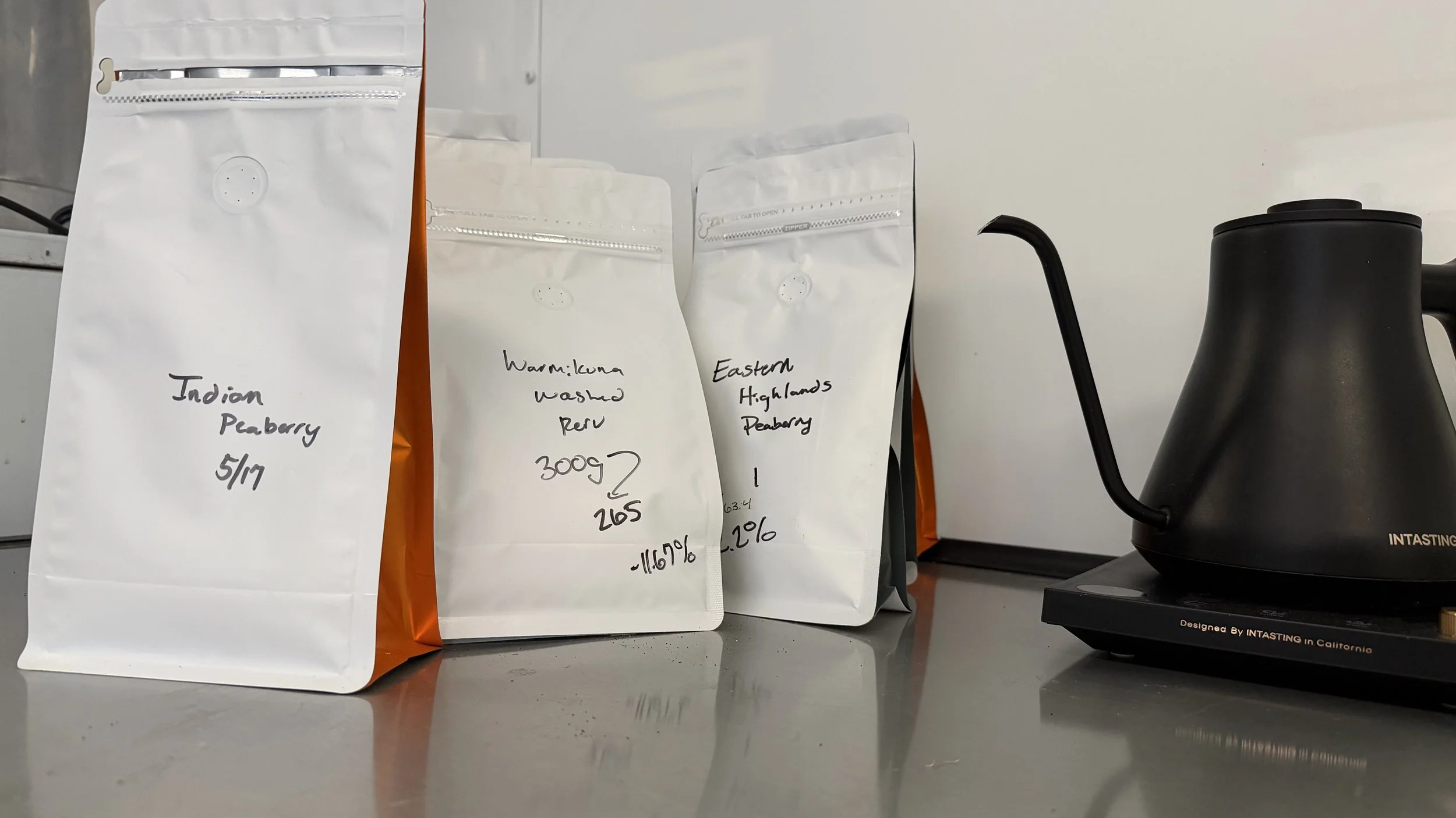 Three white resealable bags of different types of dried fruit, labeled 'Indian Peaberry,' 'Warming Luna Washed Rwu,' and 'Eastern Highlands Peaberry,' on a countertop next to a black electric kettle.