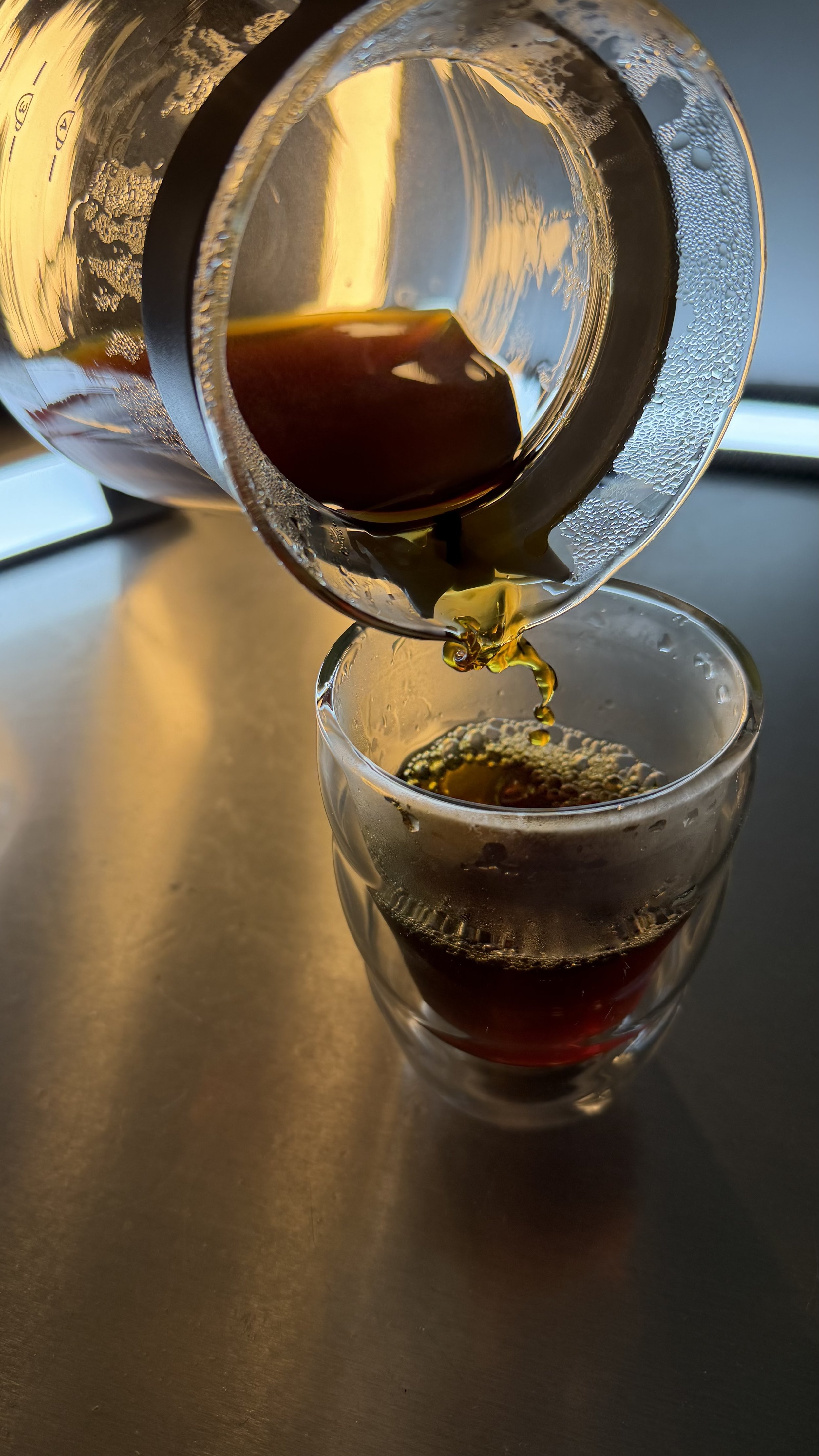 A glass of dark soda being poured from a metal can, with bubbles on the side of the can and foam forming in the glass.